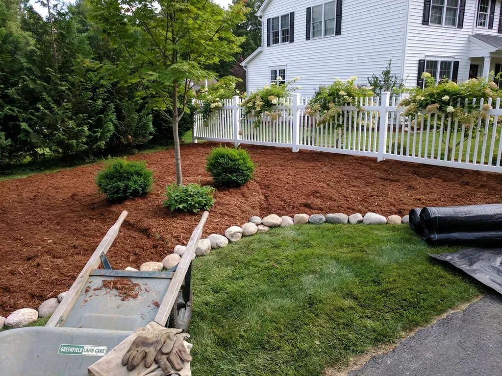 Landscaped front yard with mulch beds, shrubs, stepping stones, and white house by a fence