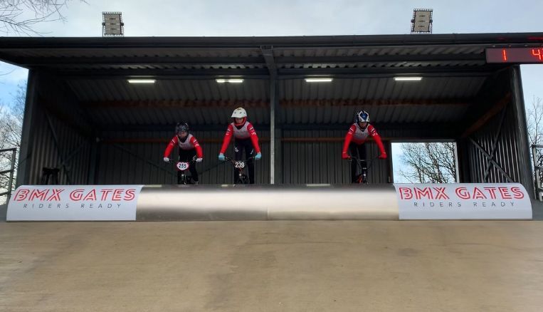 Drie BMX-rijders, in rood-zwarte shirts en met helmen op, staan ​​klaar bij de start, onder een metalen dak, klaar om te racen.