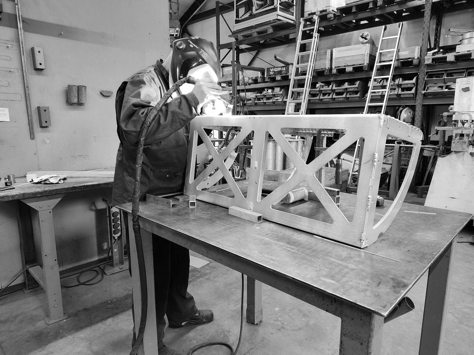 Een lasser in beschermende kleding last een metalen frame op een werkbank in een werkplaats.