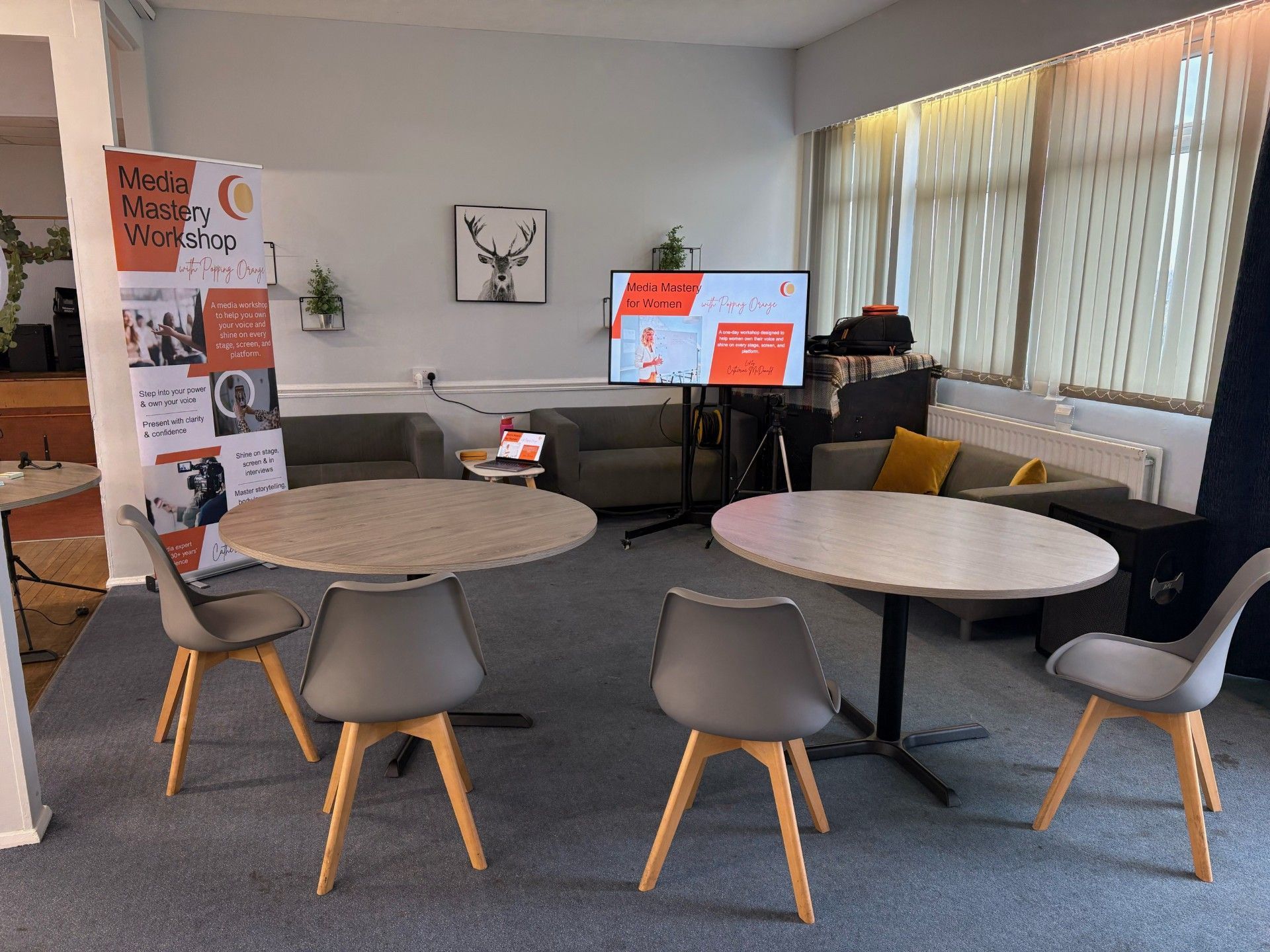 A modern office space with round tables, chairs, sofas, a TV screen, and promotional material.