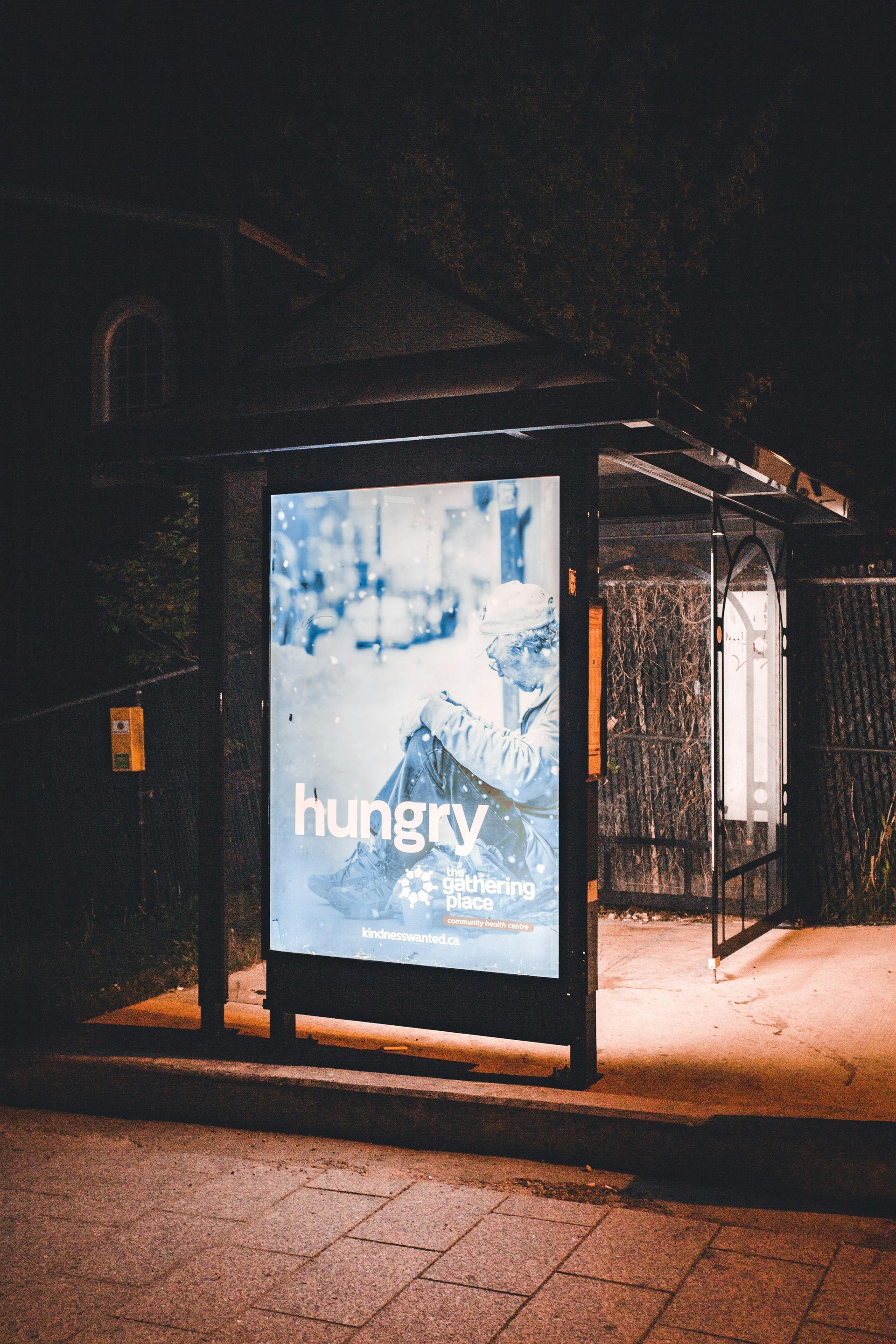Bus stop with a backlit ad that says “hungry”. Dark, nighttime setting.