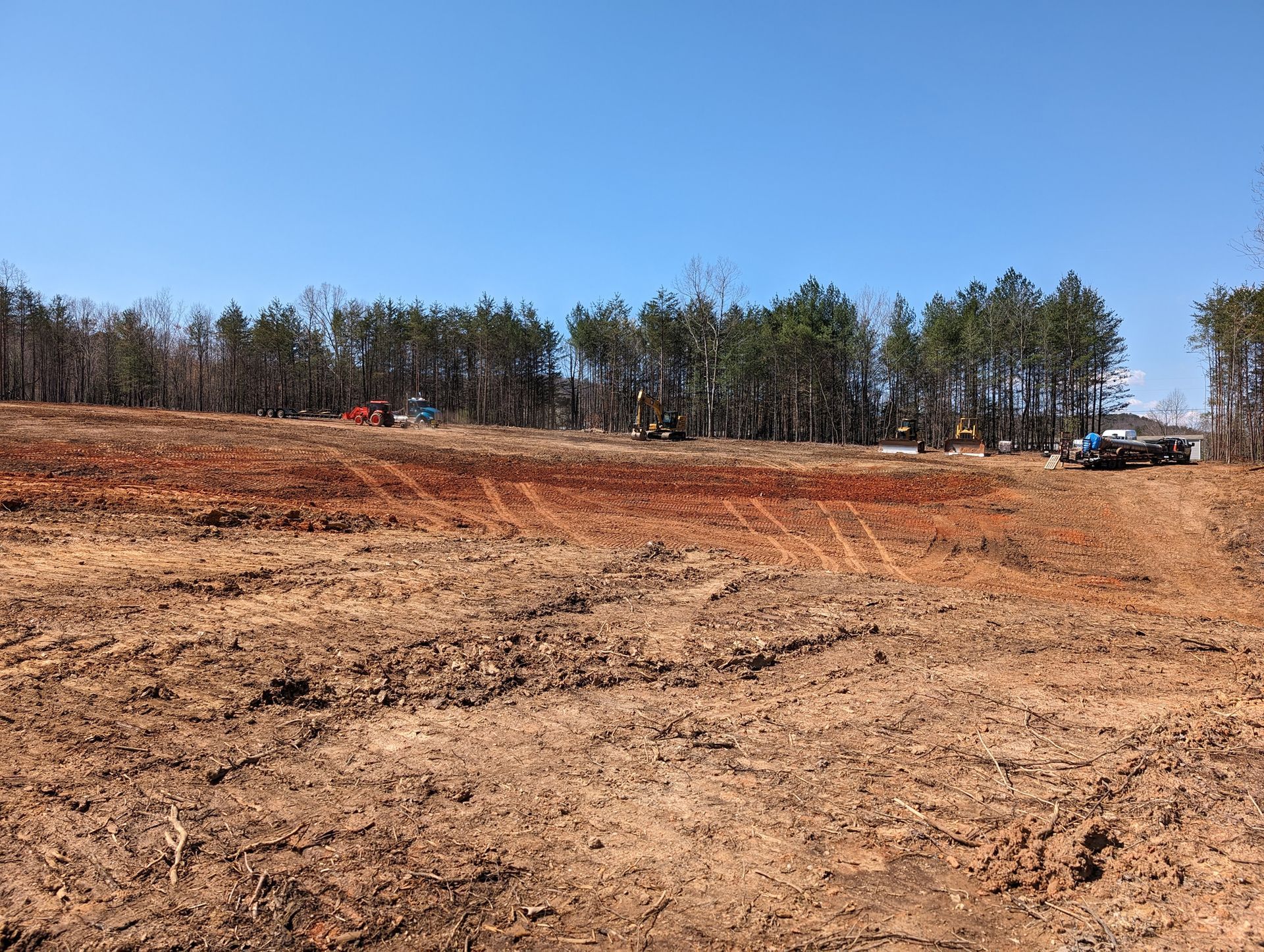 Cleared dirt lot with a tree line under a blue sky, and a small construction vehicle at right