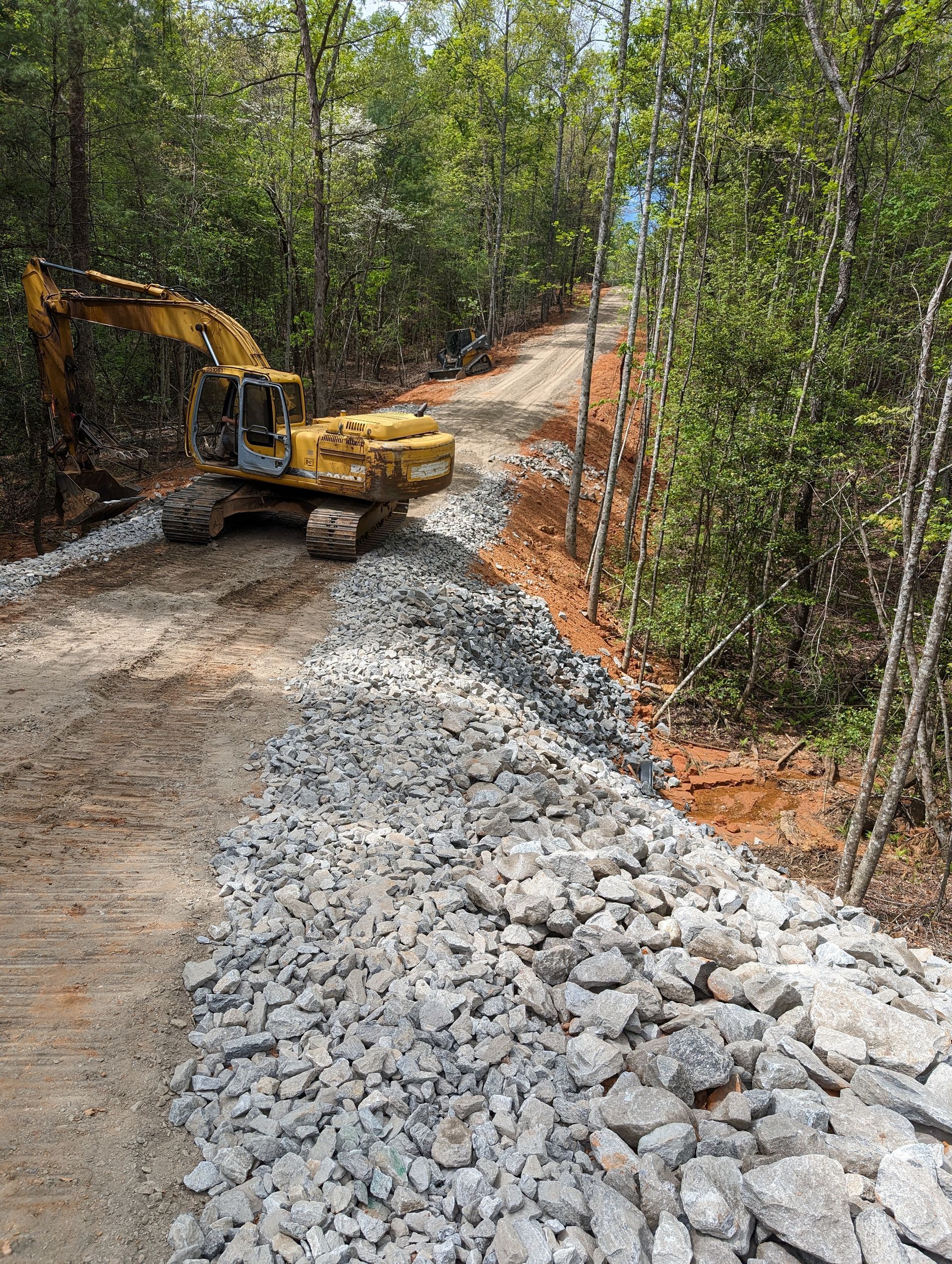 Excavator grading a gravel road through a wooded area, with a pile of rocks in the foreground.
