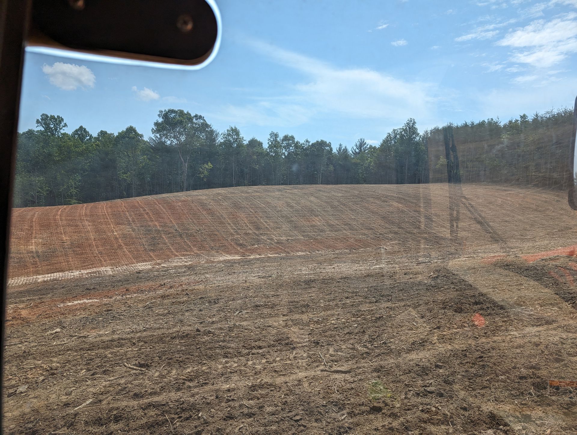 Rough dirt field under blue sky, with a tree line in the distance and a car windshield edge at top left
