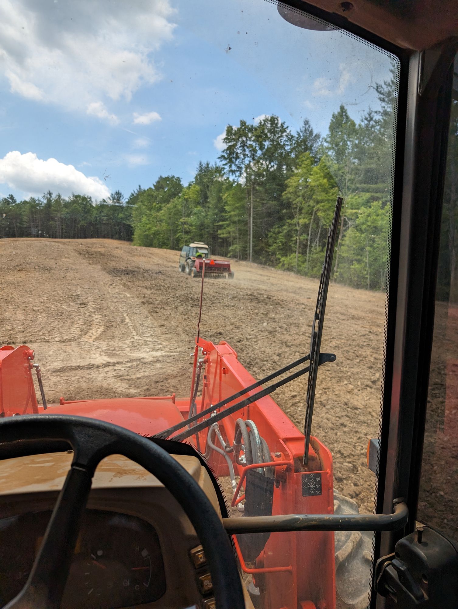 View from inside an orange excavator cab at a dirt worksite, with another machine in the distance.