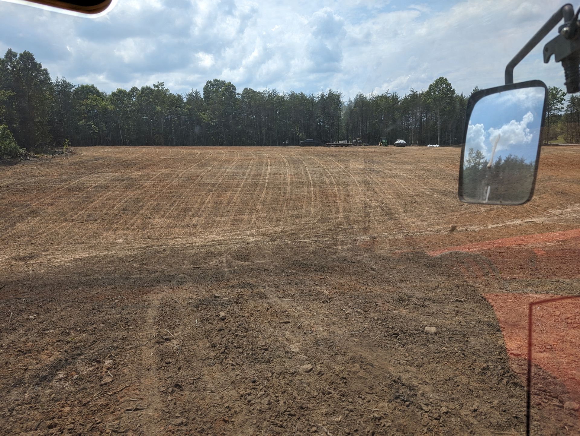 Wide dirt field under cloudy sky, with trees in the distance and a vehicle mirror visible on the right.