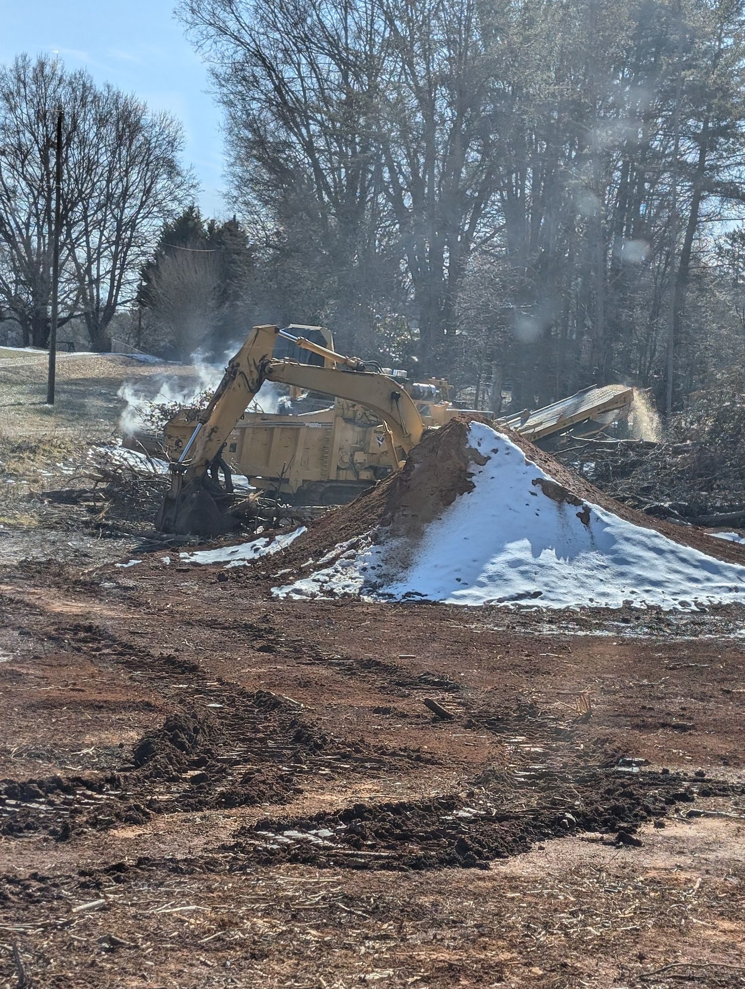 Yellow excavator clearing dirt beside a snowy mound in a wooded field.