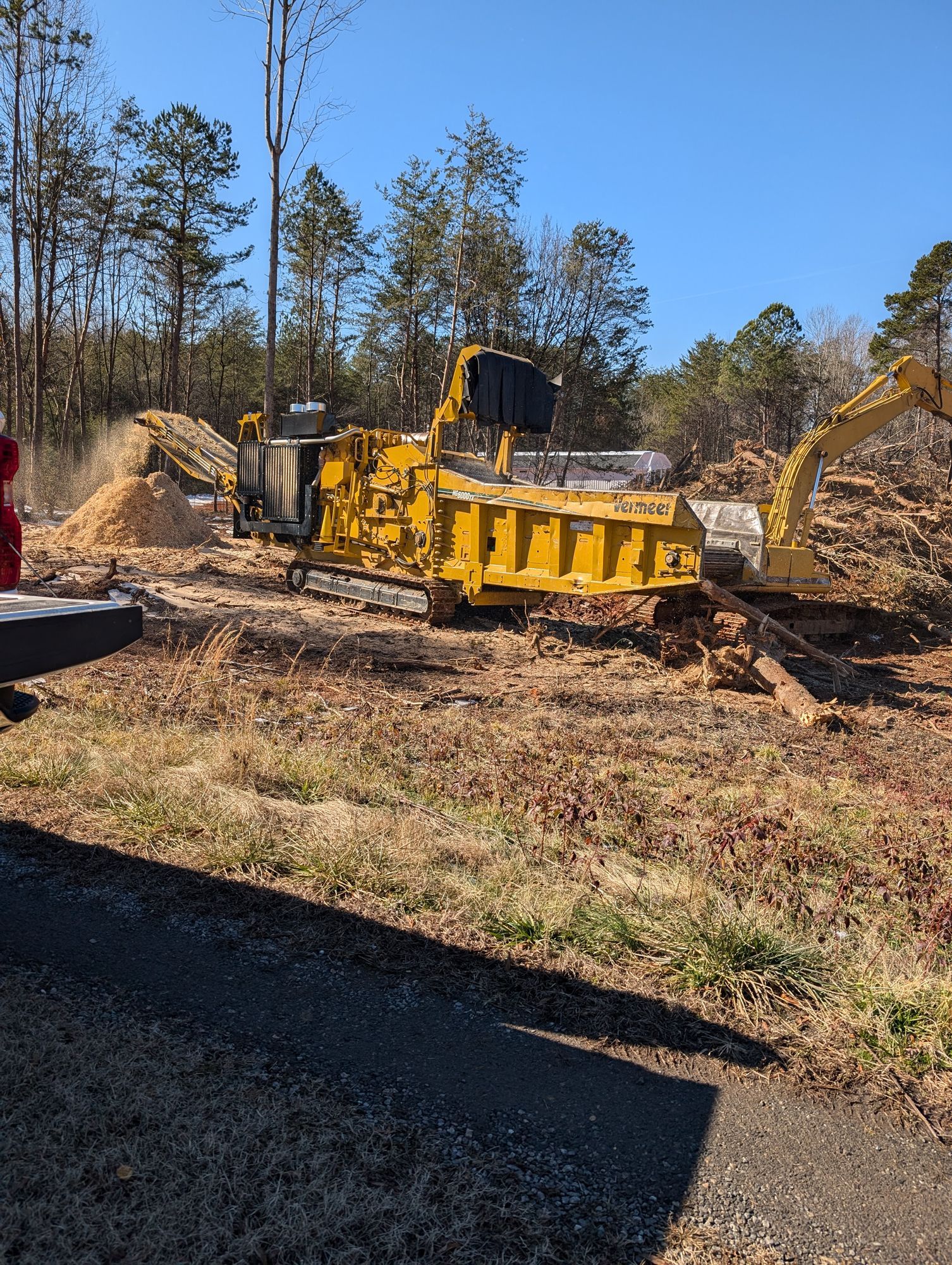 Yellow industrial wood chipper in a wooded clearing, with a worker nearby and piles of brush around it.