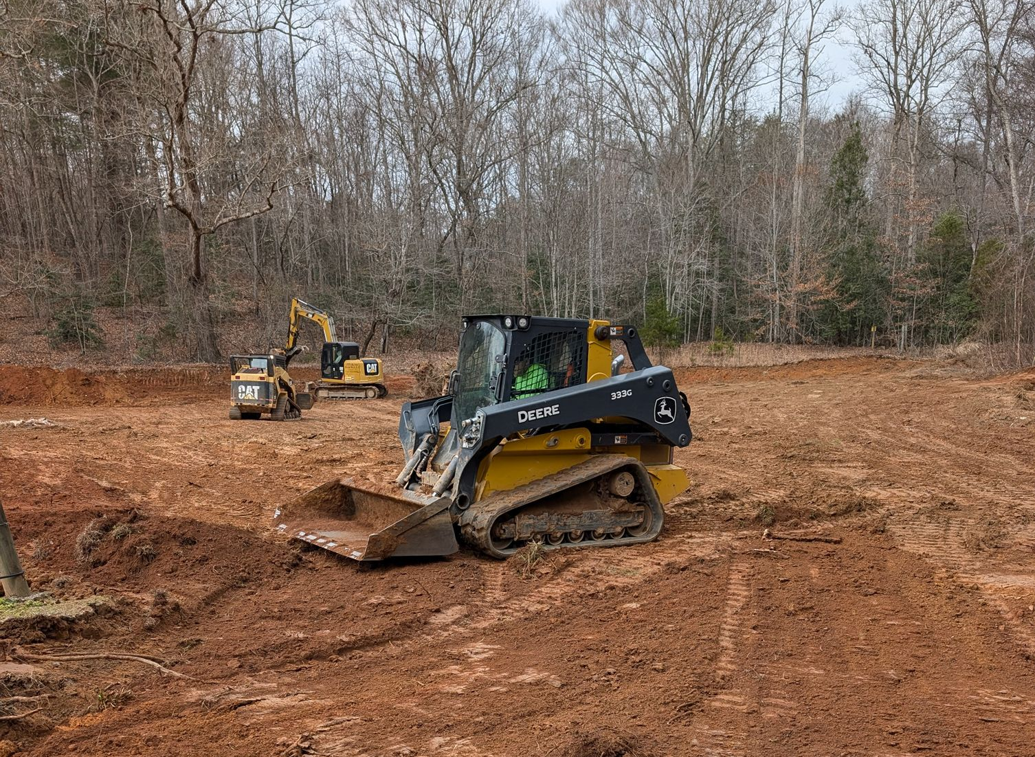 Two construction machines on a dirt lot with leafless trees in the background