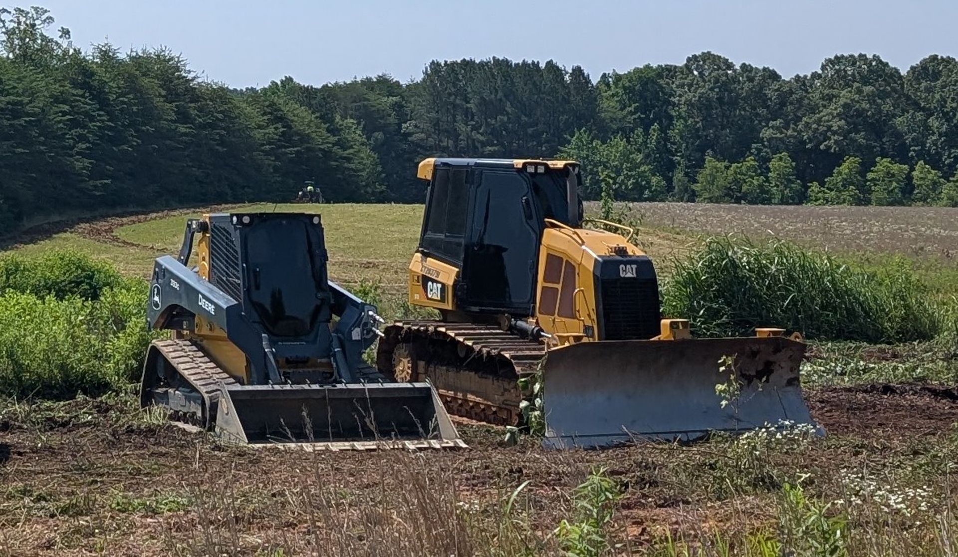 Two bulldozers clearing a field, one gray and one yellow, with trees and crops in the background