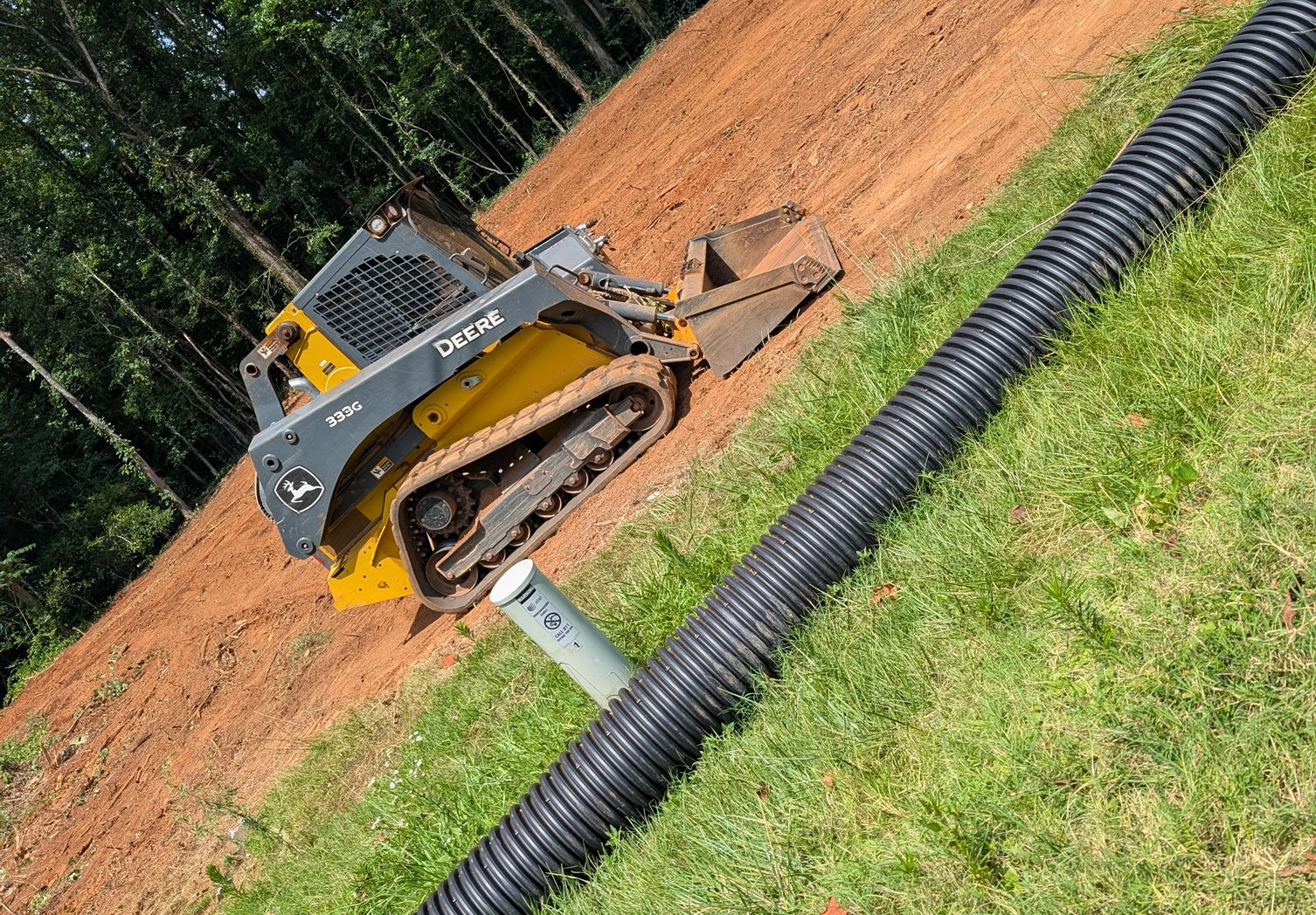 Yellow skid steer on a dirt slope beside a black corrugated drainage pipe and grassy edge.