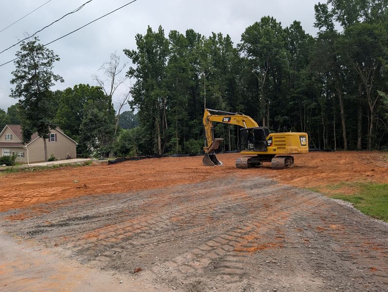 Yellow excavator on a dirt lot with trees and a house in the background