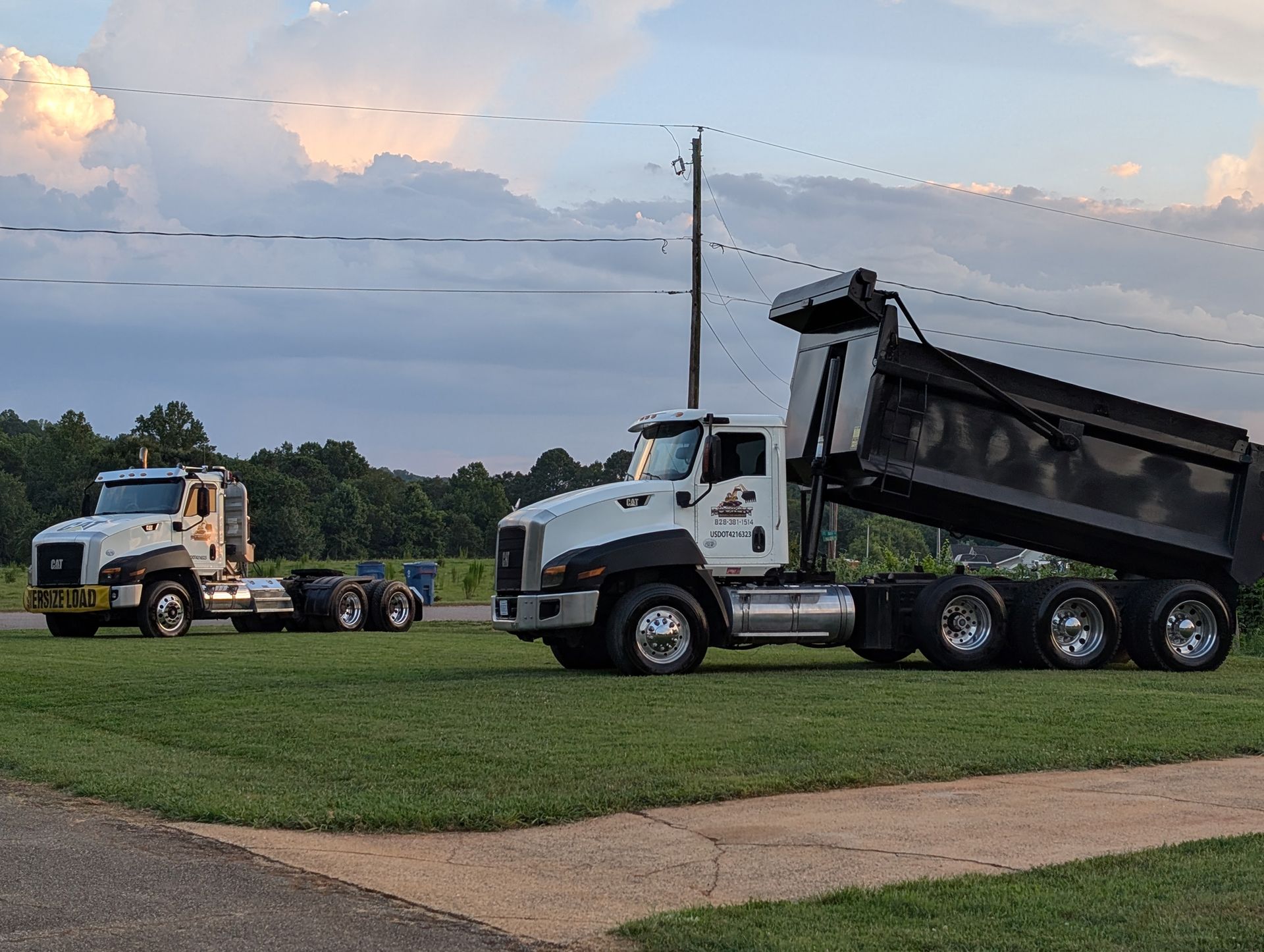 Two dump trucks parked on grass near a road at dusk, one with its bed raised.
