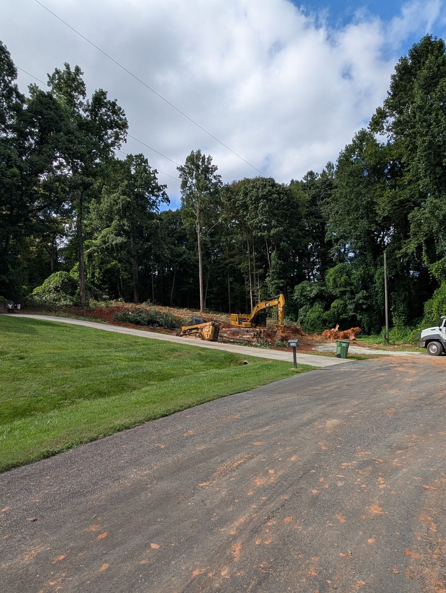 Gravel driveway beside a wooded lot with cleared trees, construction equipment, and a partly cloudy sky