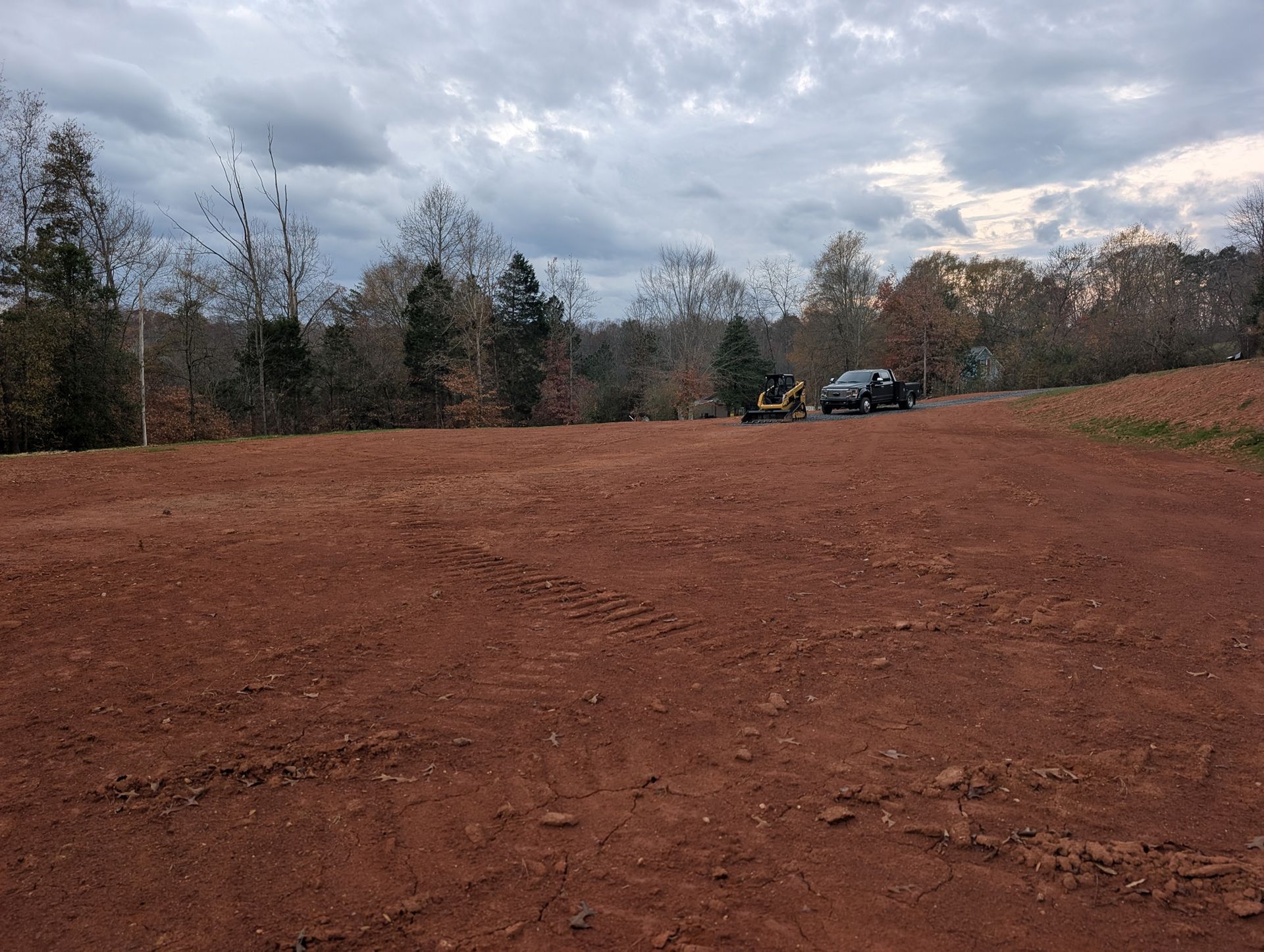 Dirt clearing beside a tree line under a cloudy sky, with a vehicle in the distance.