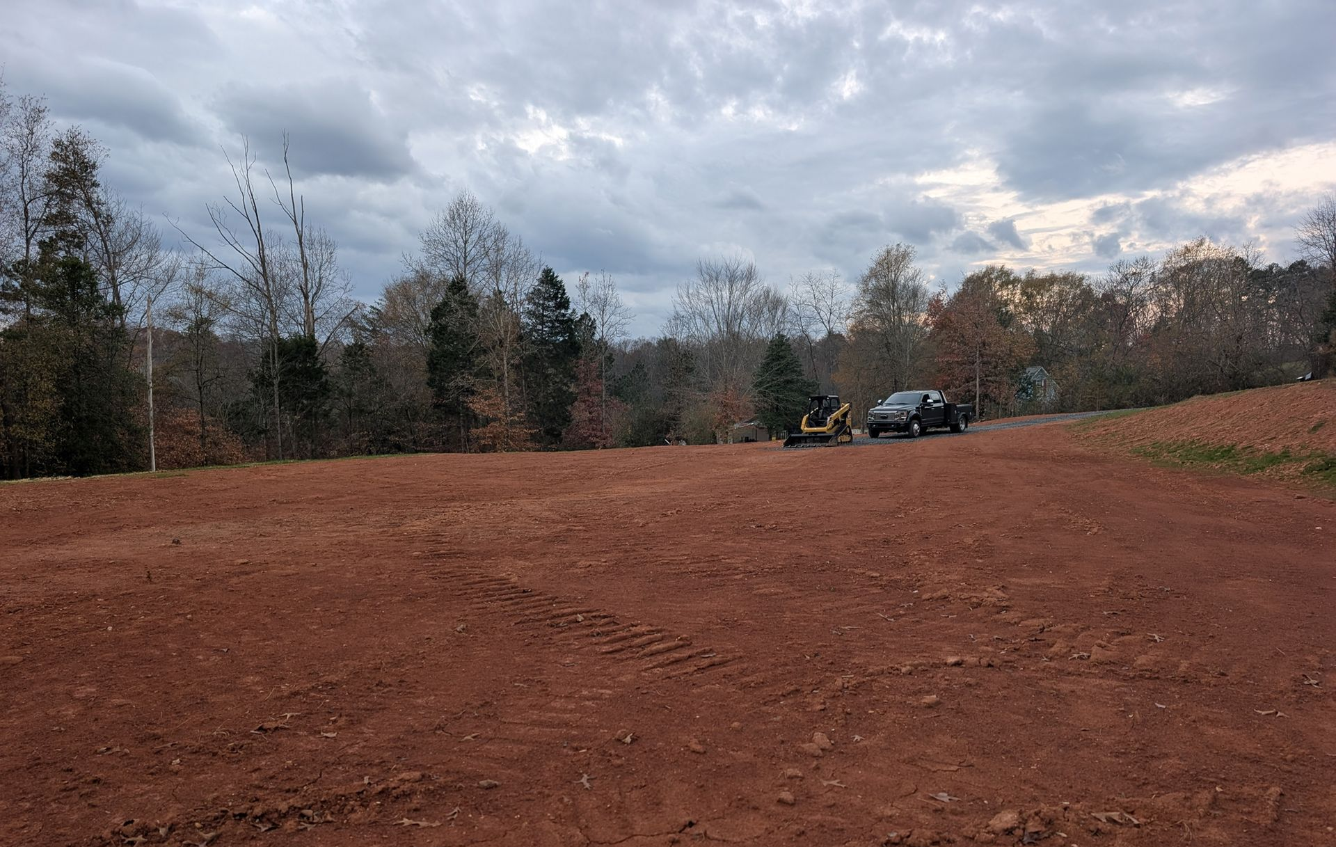 Red dirt lot with trees and cloudy sky, a parked vehicle in the distance