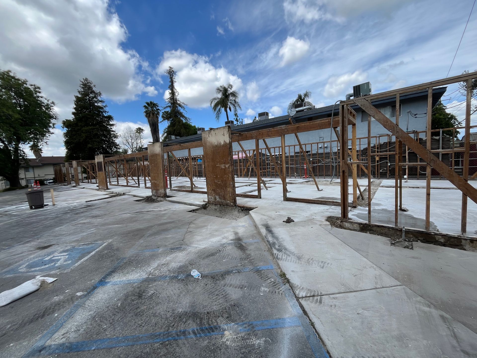 A construction site featuring a wooden frame structure on a concrete slab in a parking lot under a cloudy blue sky.