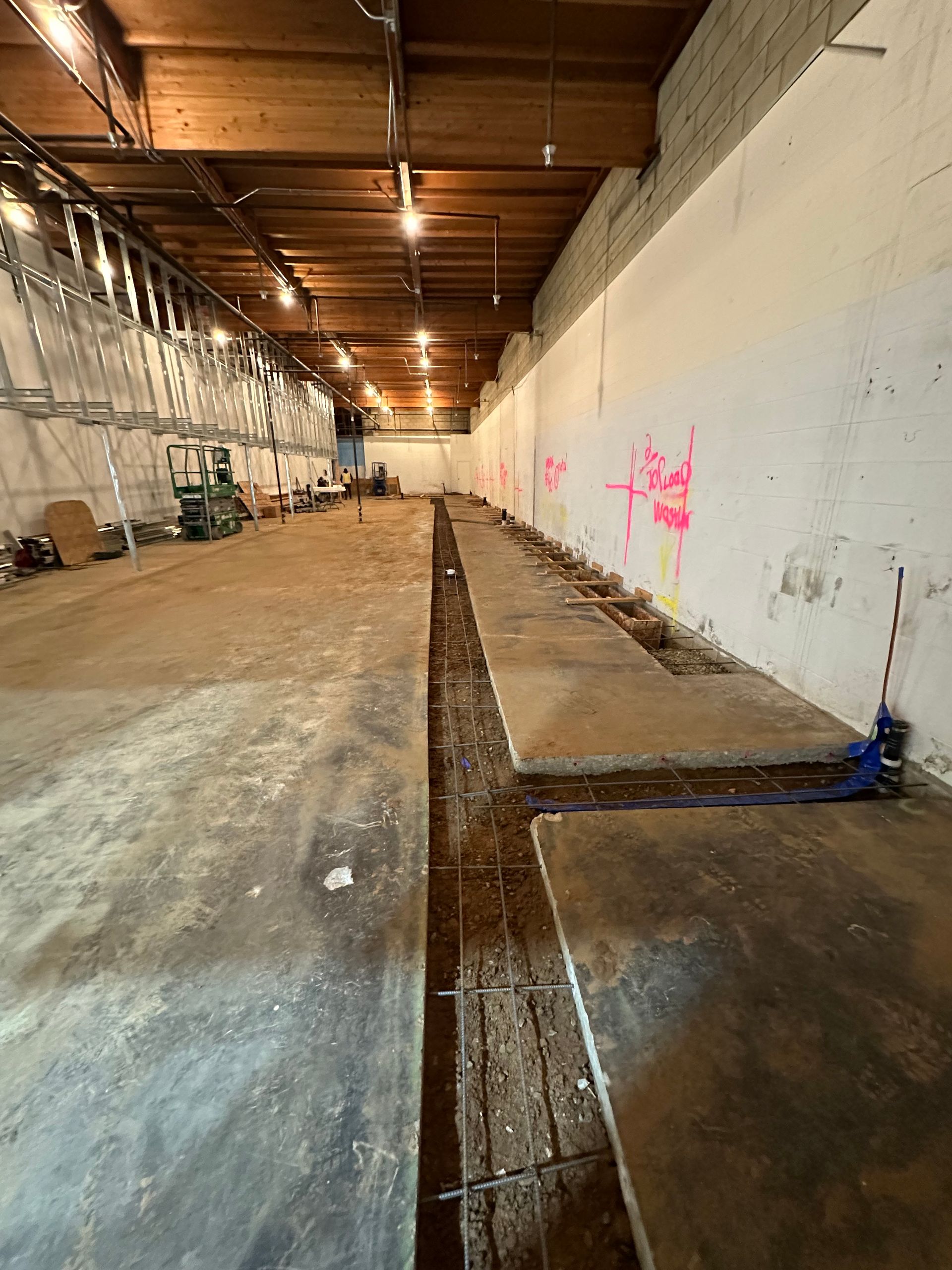 Interior construction site with a long concrete trench, exposed rebar, and a white wall with pink graffiti markings.