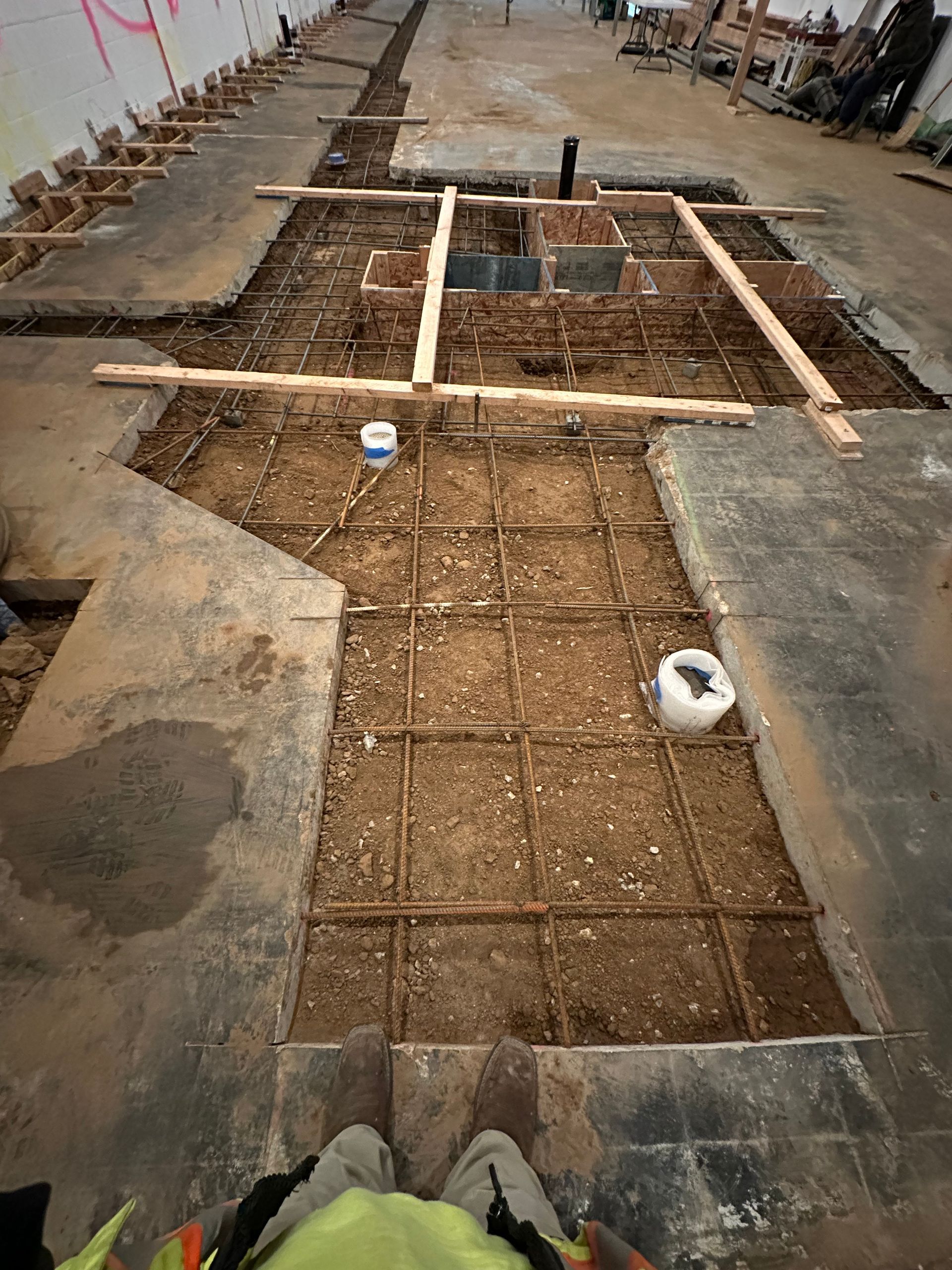 An indoor construction site showing a dirt-filled floor trench reinforced with a metal rebar grid and wooden frame forms.