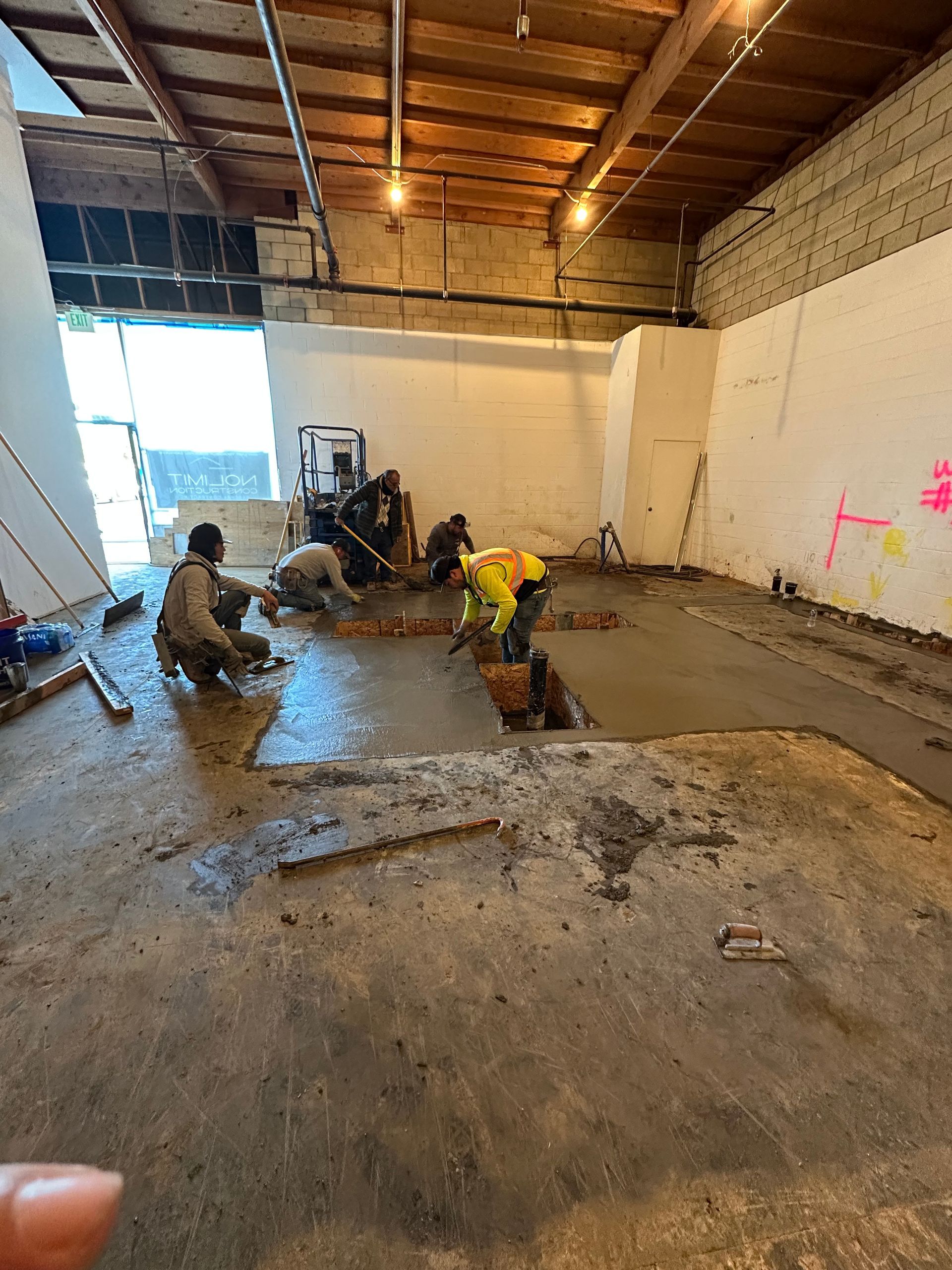 Construction workers finish laying a new concrete floor section inside an industrial building with exposed wood rafters.