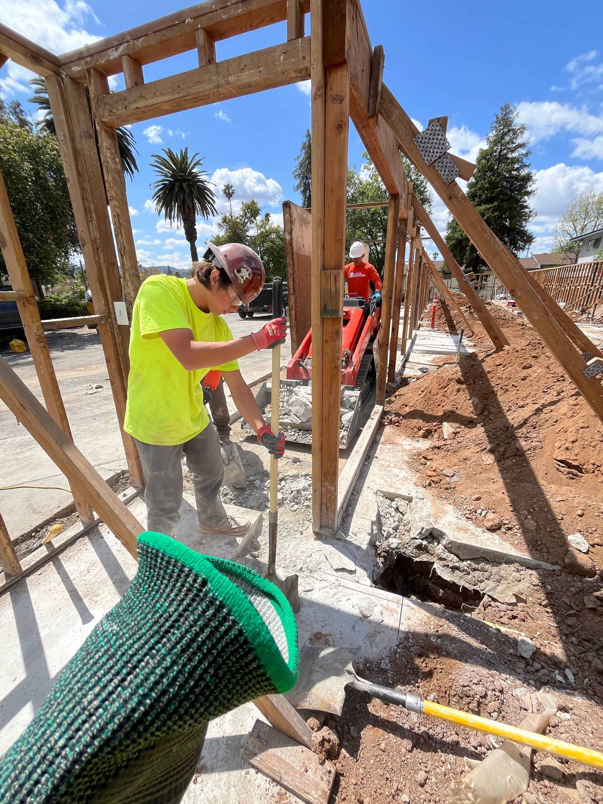 Construction workers in safety gear digging and clearing dirt near a wooden frame structure on a sunny day.