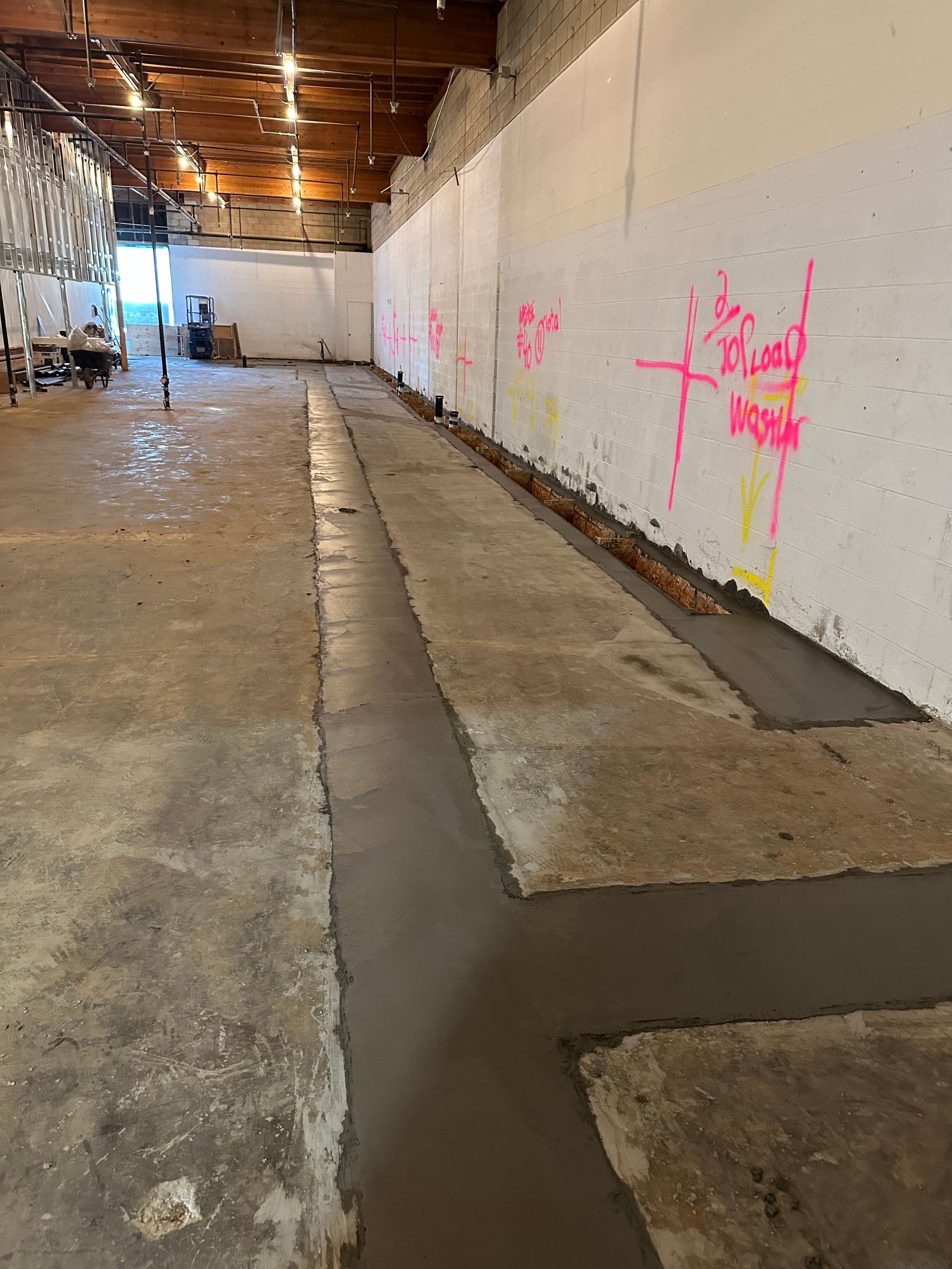 An unfinished indoor construction site with freshly poured concrete channels along a white wall marked with neon spray paint.
