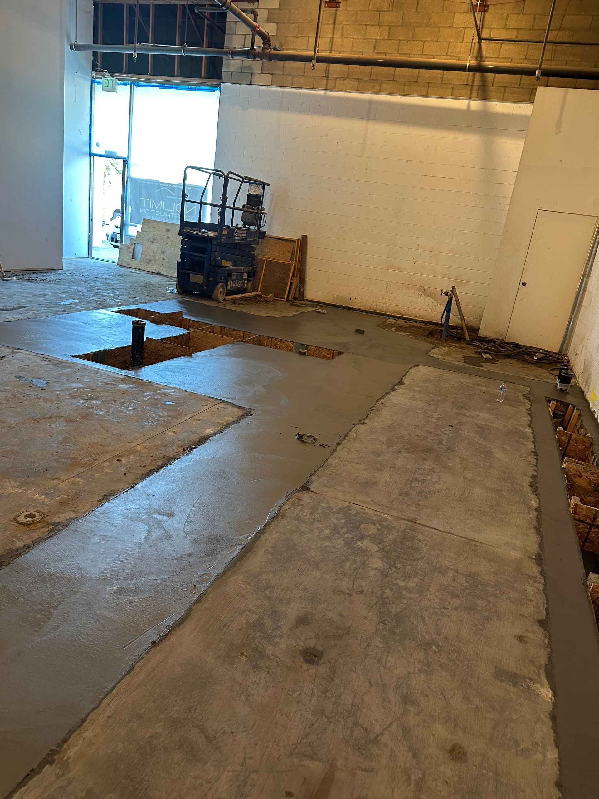 An indoor construction site featuring a concrete floor with open trenches, exposed walls, and a scissor lift.