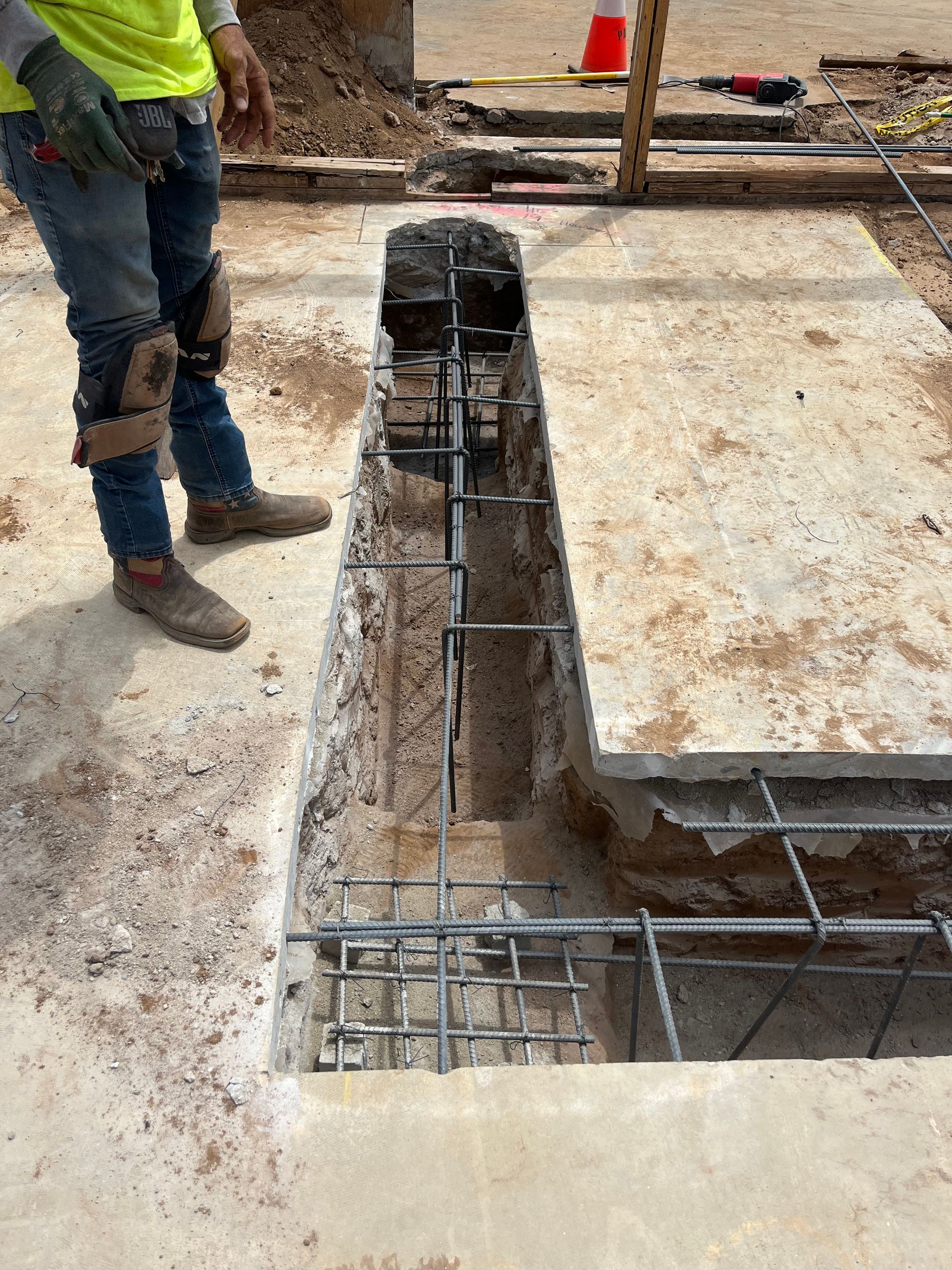 A construction worker standing by a rectangular trench in a concrete slab, revealing steel rebar reinforcement.