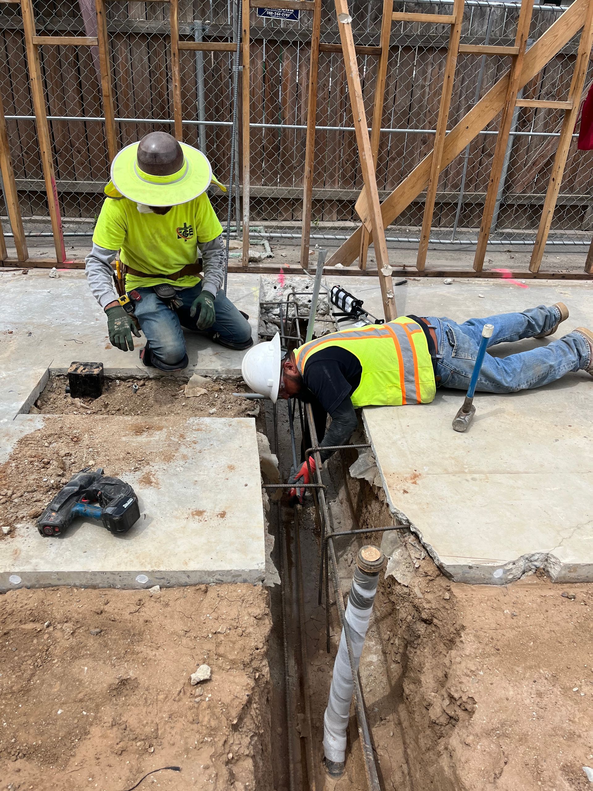 Two construction workers in high-visibility vests work in a trench at a building site, one kneeling and one lying prone.