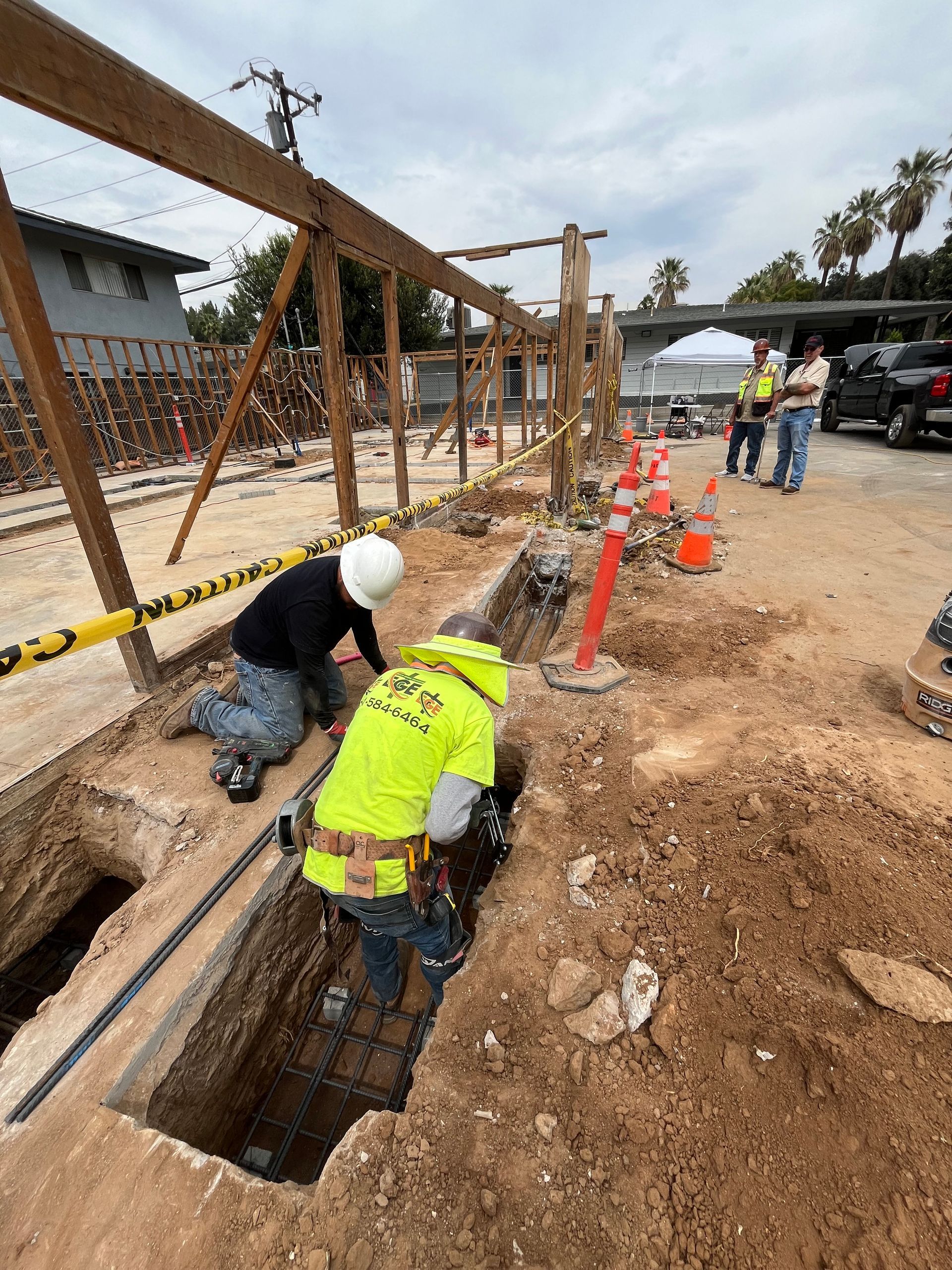 Construction workers in safety gear work in a trench at an outdoor site with wooden framing nearby.