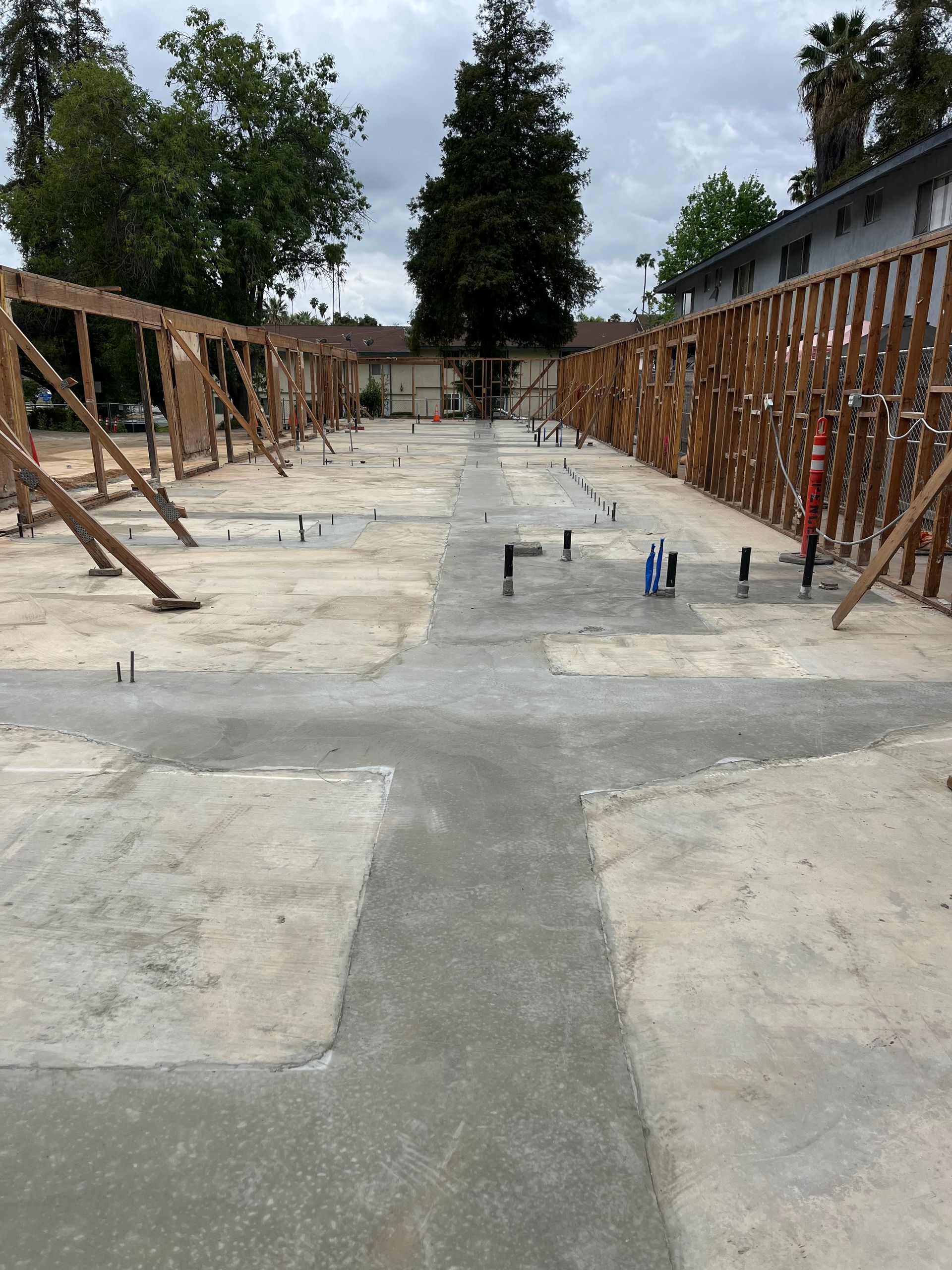 Construction site featuring a concrete slab foundation with plumbing pipes and wooden wall frames under an overcast sky.