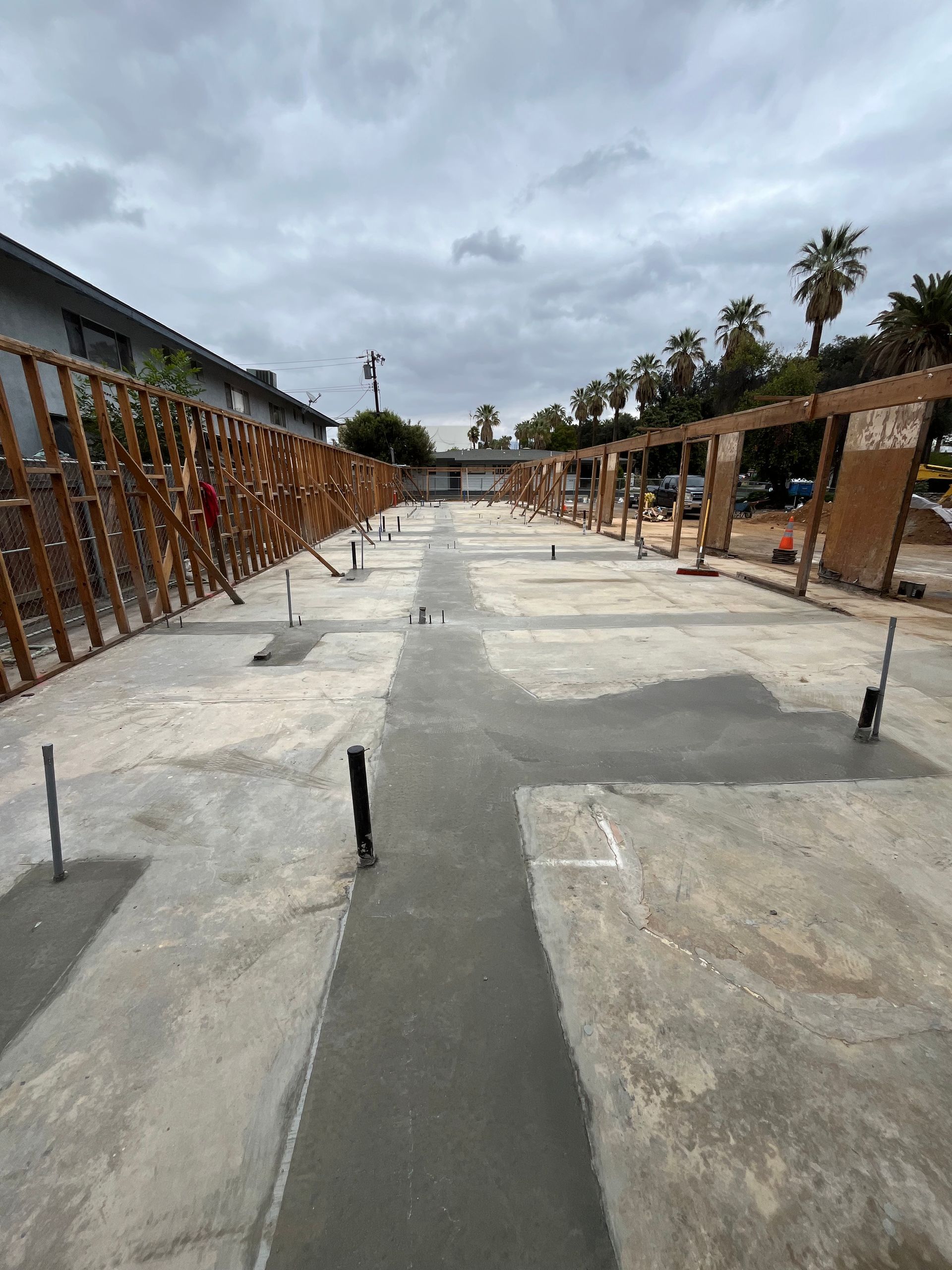A construction site showing a concrete foundation, plumbing pipes, and wooden framing walls under a cloudy sky.