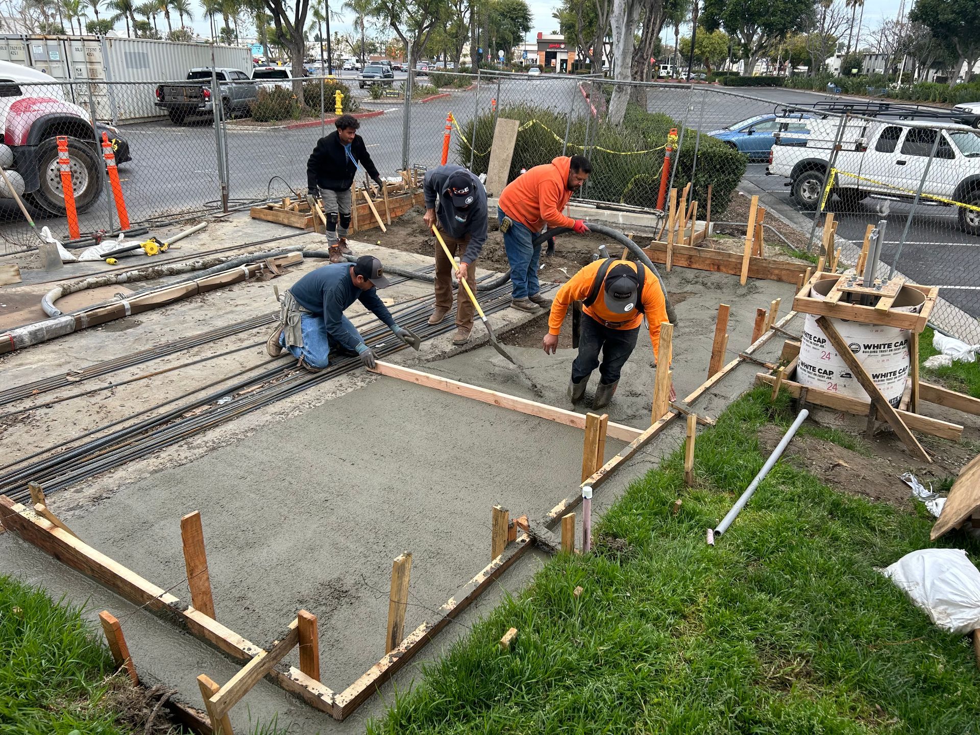 Workers in bright orange and blue shirts smoothing freshly poured wet concrete into wooden forms at a construction site.