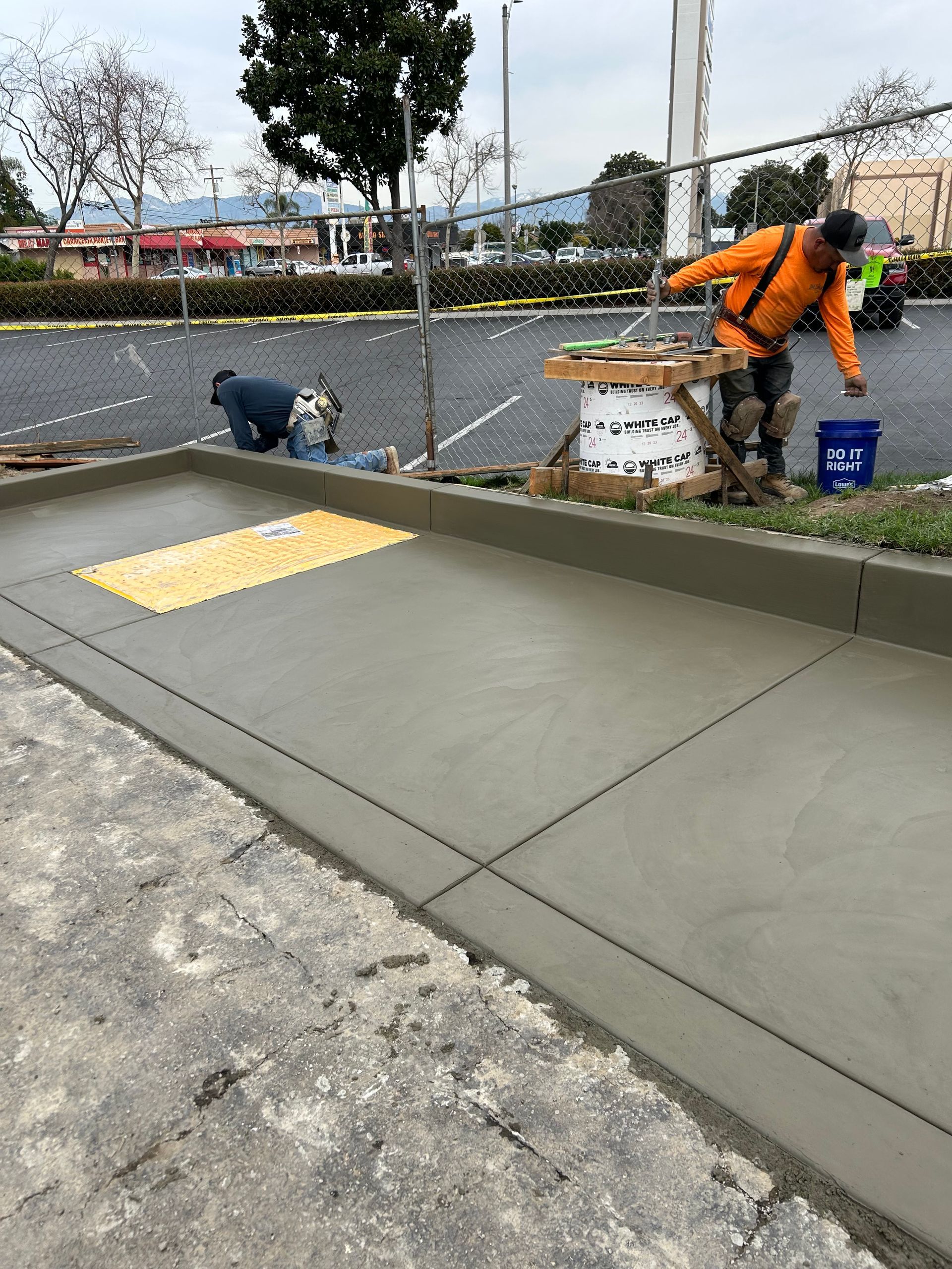 Two construction workers finish a newly poured concrete sidewalk with a yellow tactile paving mat.