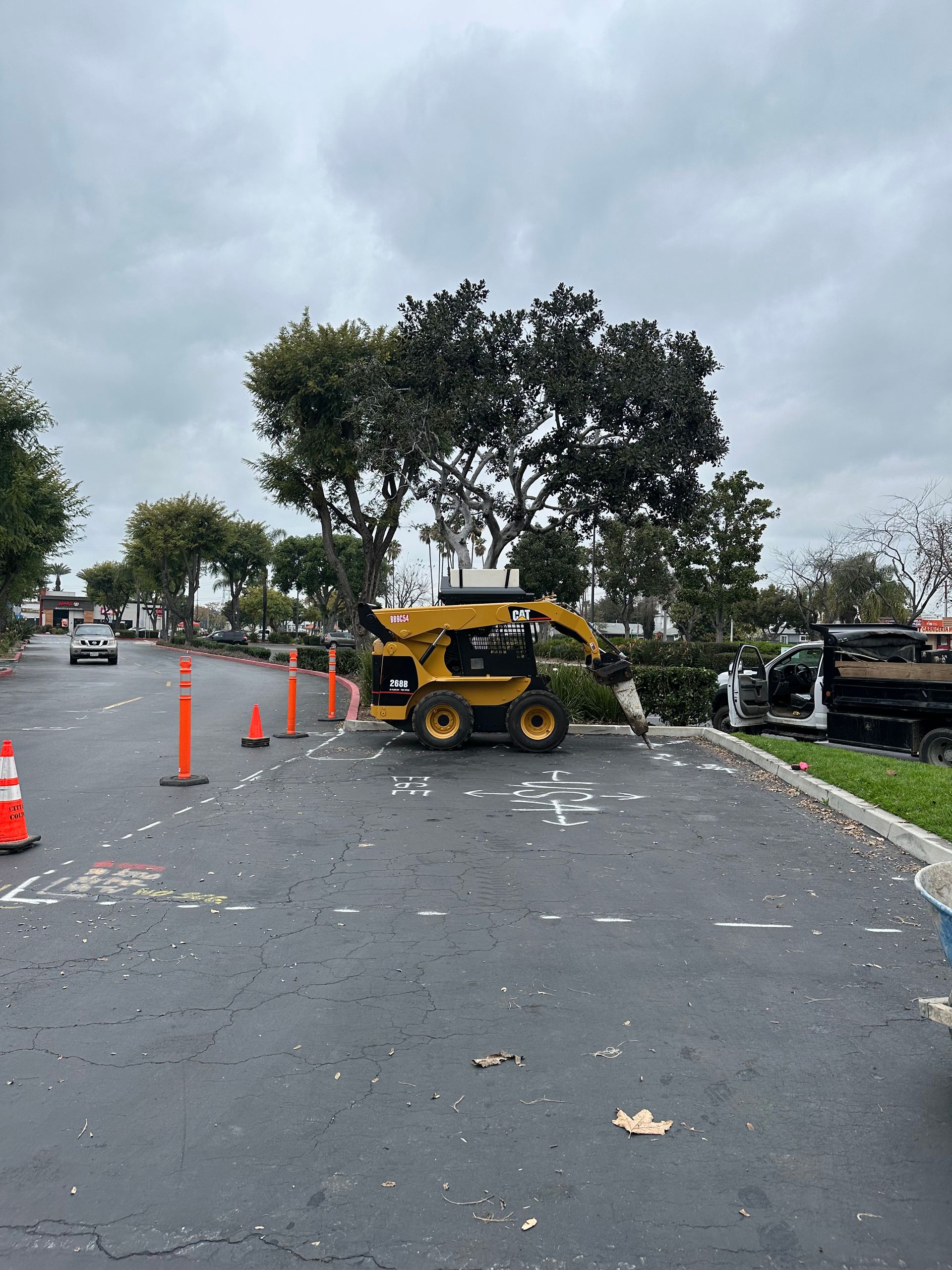 A yellow skid-steer loader with a hydraulic hammer attachment sits in a parking lot marked with construction spray paint.