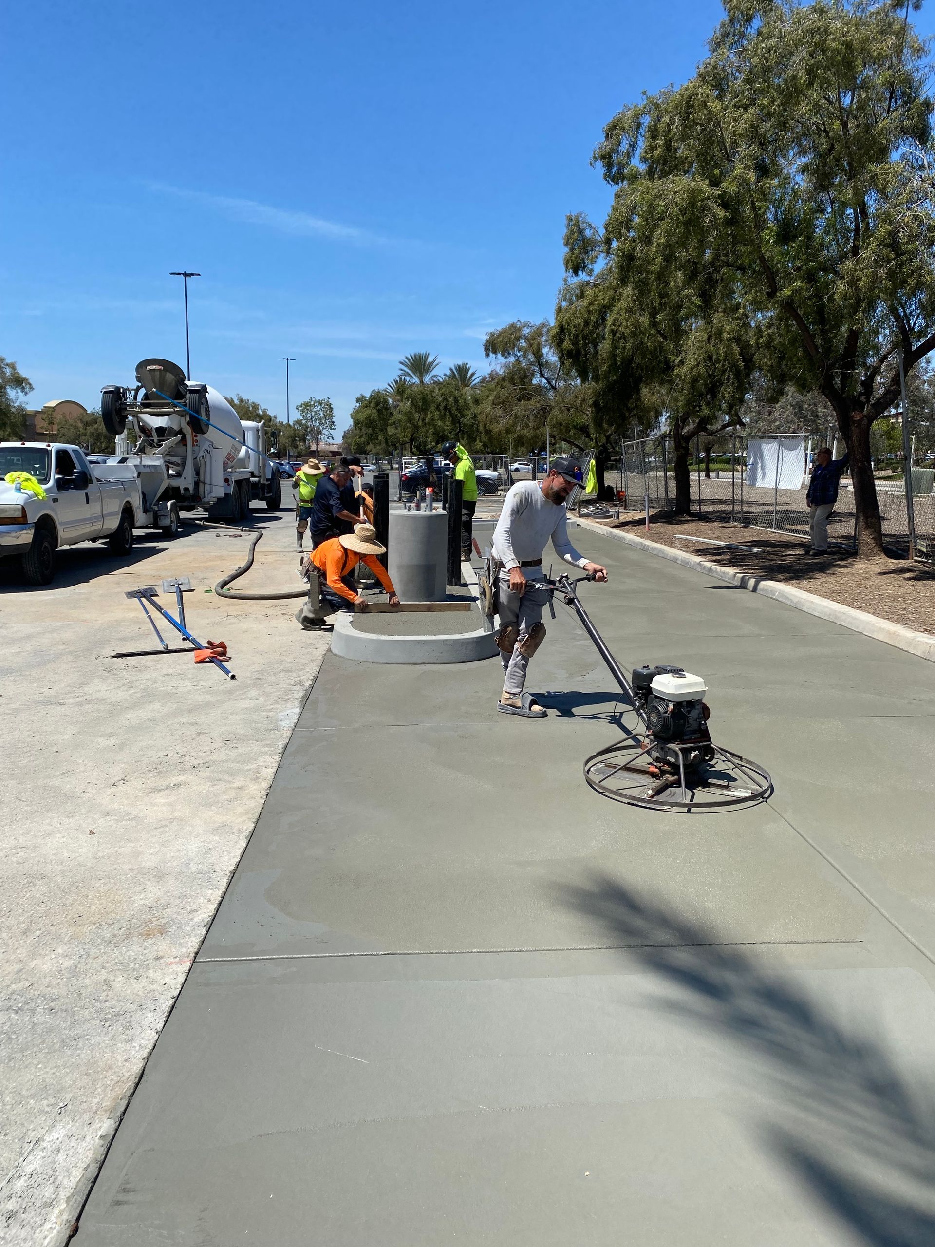 Construction workers smooth wet concrete on a new parking lot path with a power trowel near a parked cement truck.