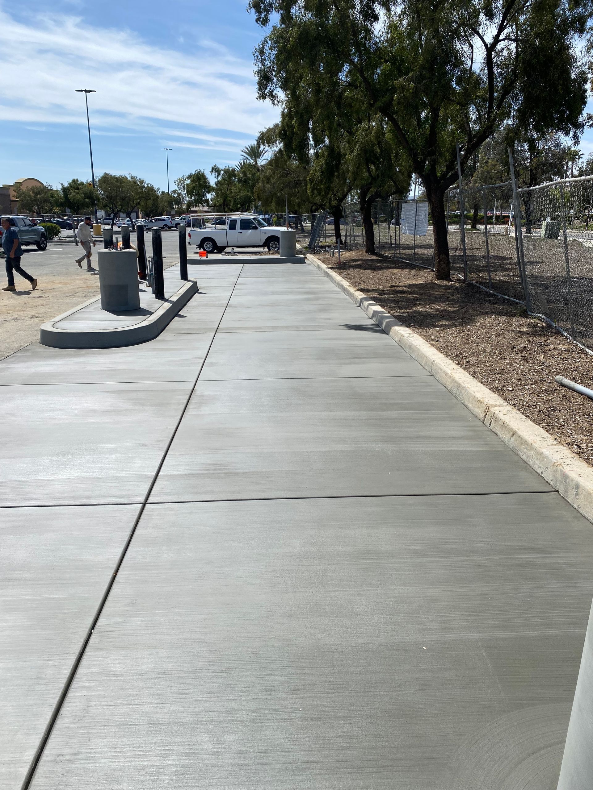 A new grey concrete sidewalk bordered by a low curb, landscaping, and bollards in a parking area under a bright sky.