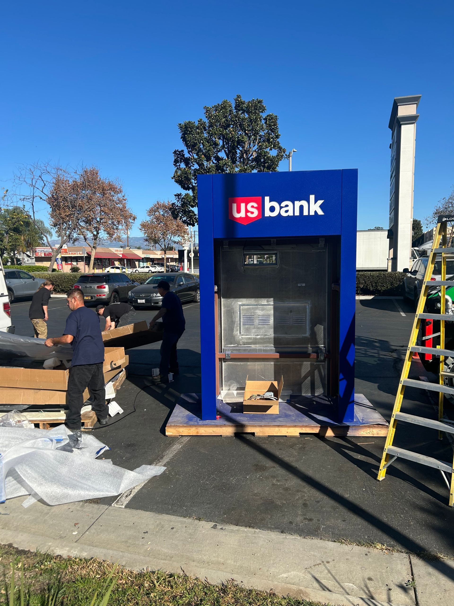 Workers installing a new U.S. Bank ATM kiosk in an outdoor parking lot on a sunny day.