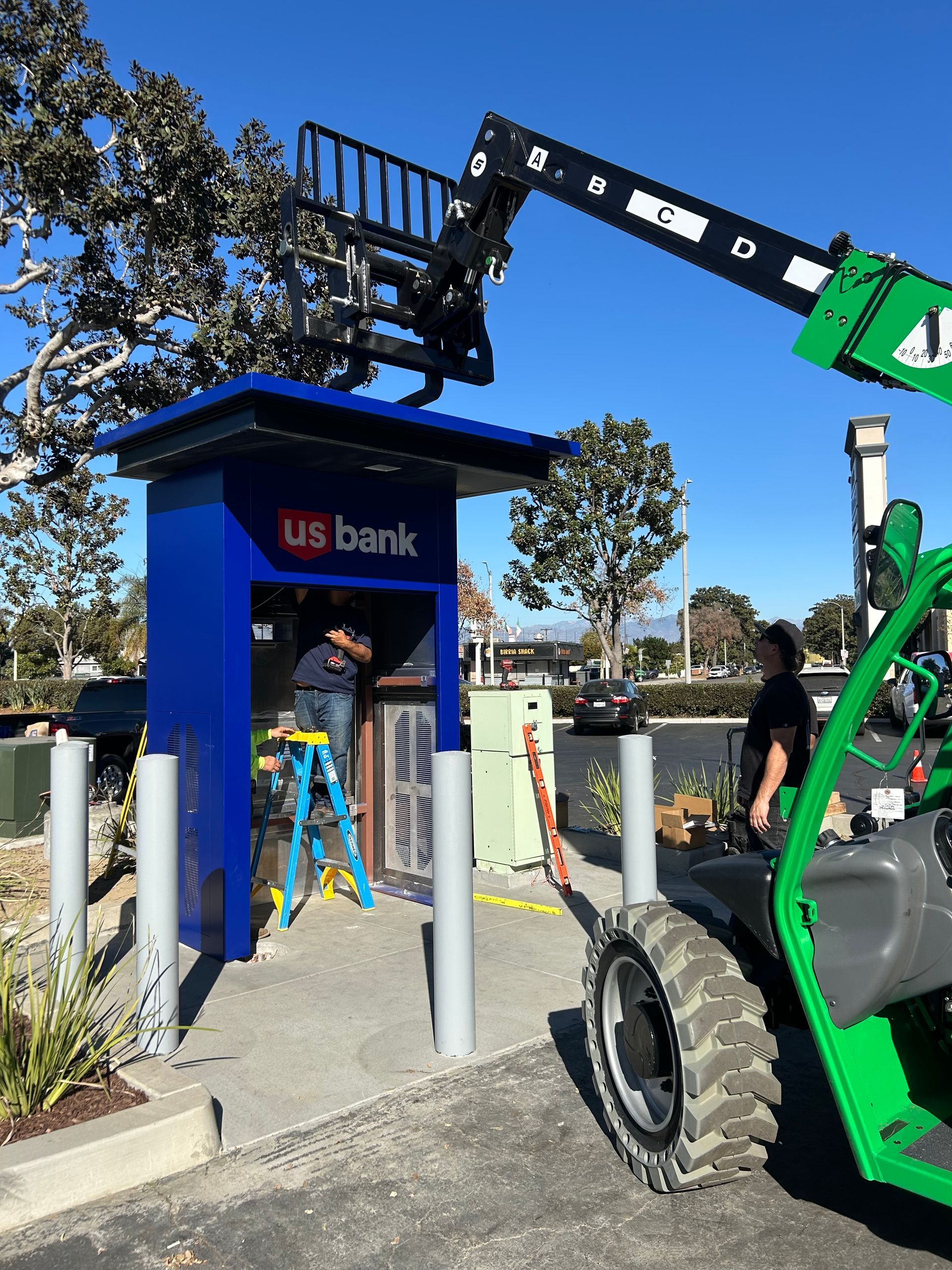 Workers install a blue U.S. Bank ATM structure using a green construction telehandler in a sunny outdoor lot.