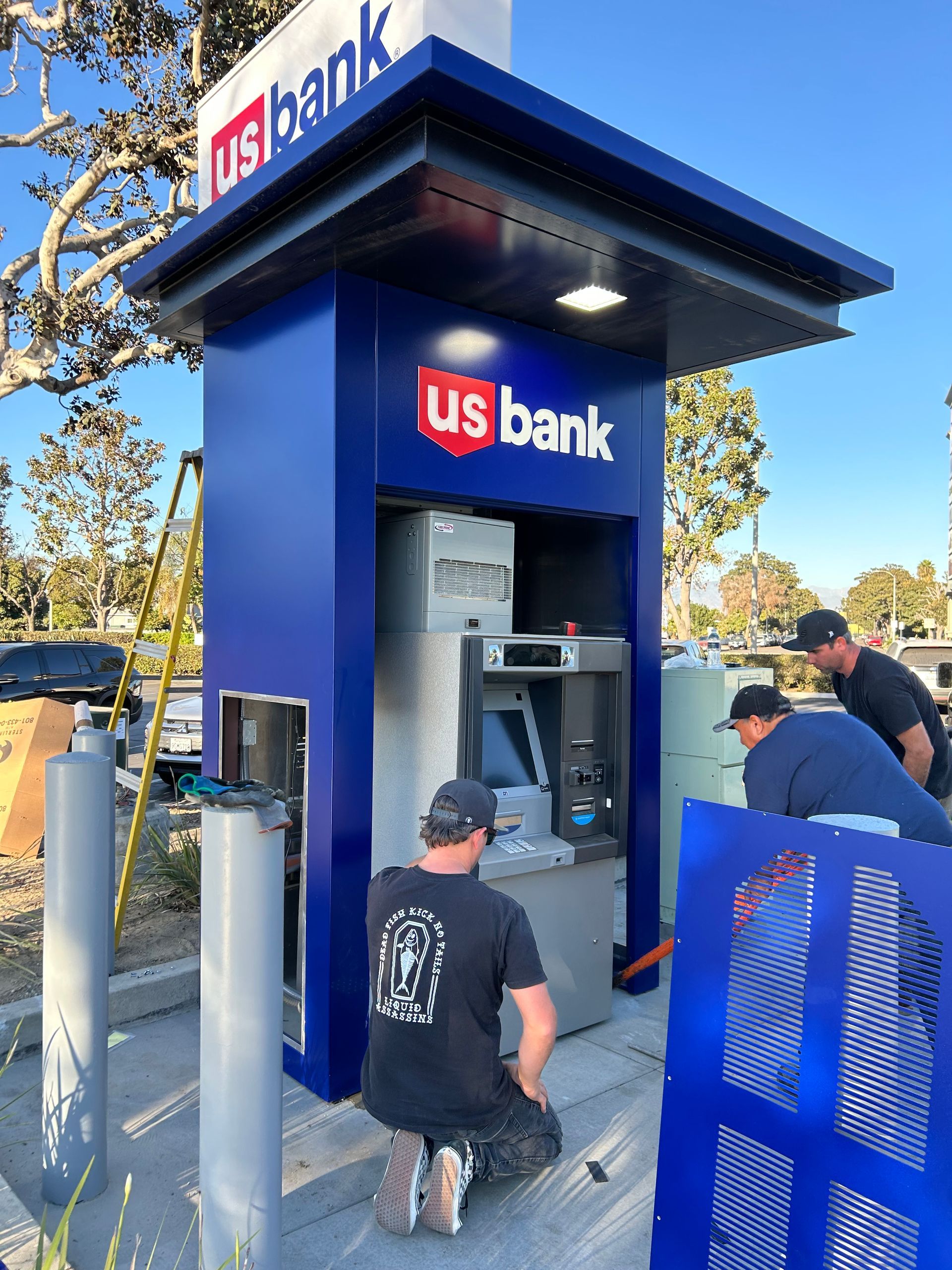 Three people work on a U.S. Bank ATM installation outdoors under a bright blue sky.