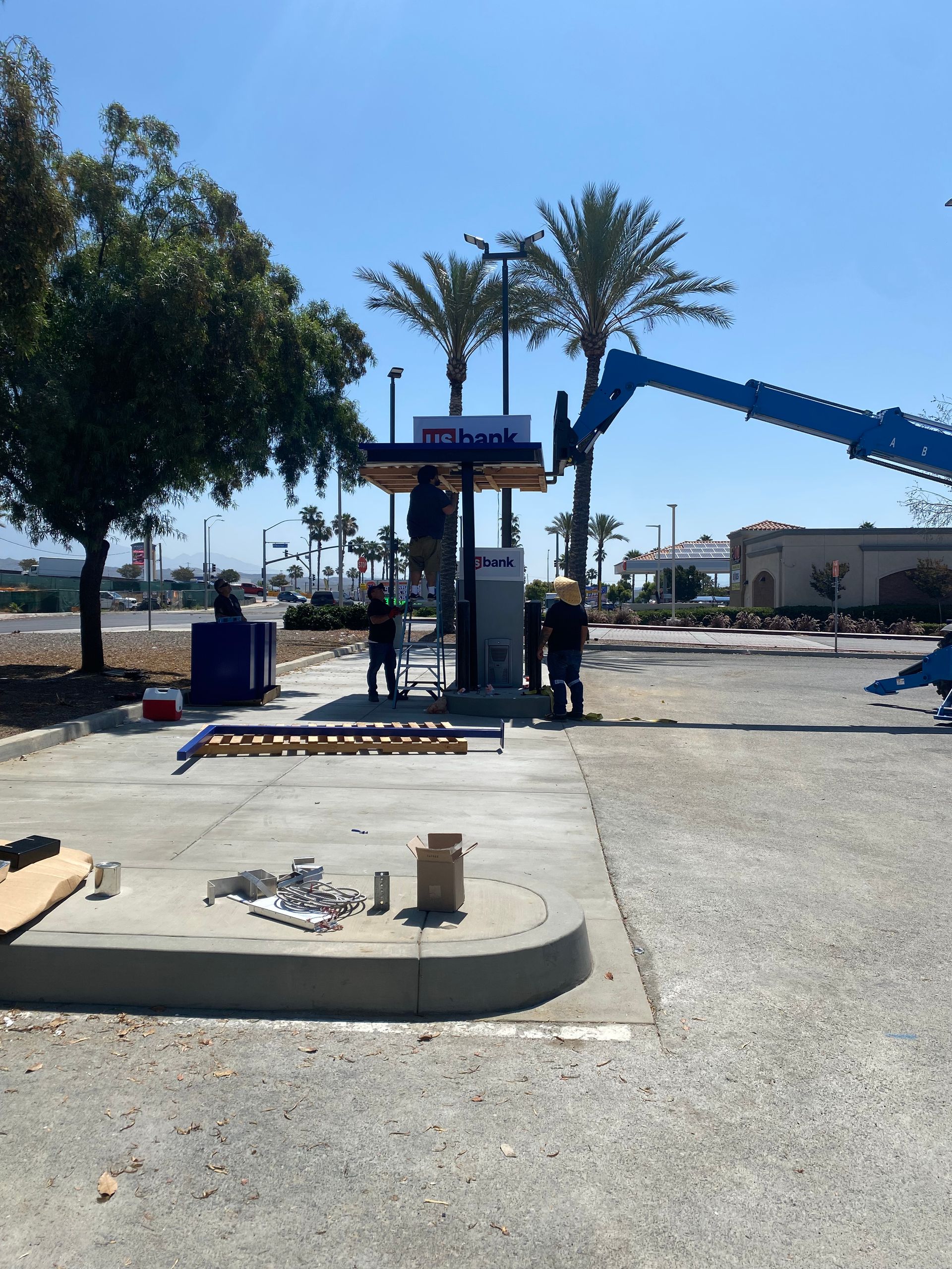 Workers installing a sign atop a covered structure in an outdoor parking lot with palm trees in the background.