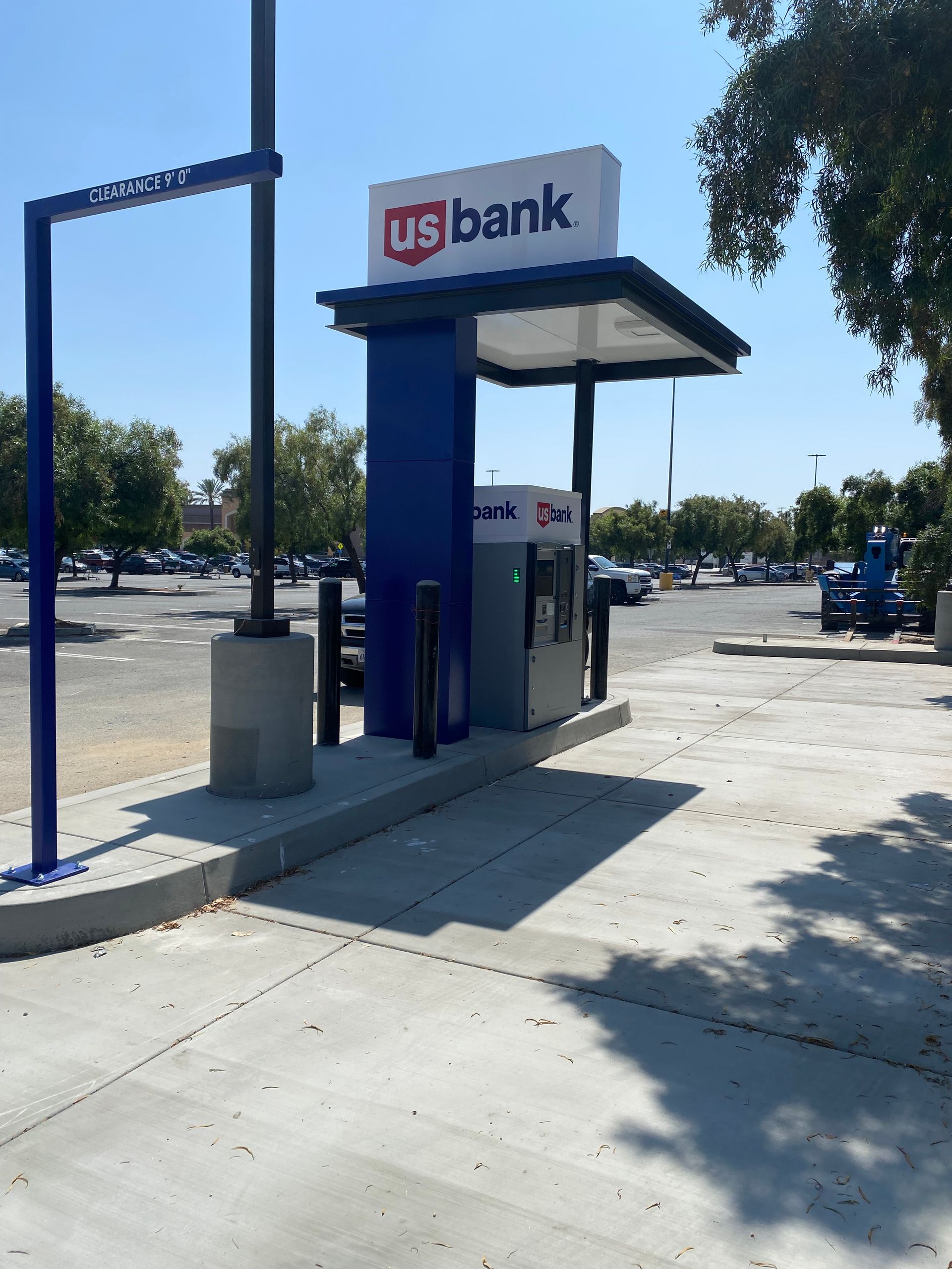 A U.S. Bank ATM stand with a blue and white canopy, situated in a paved parking lot on a sunny day.