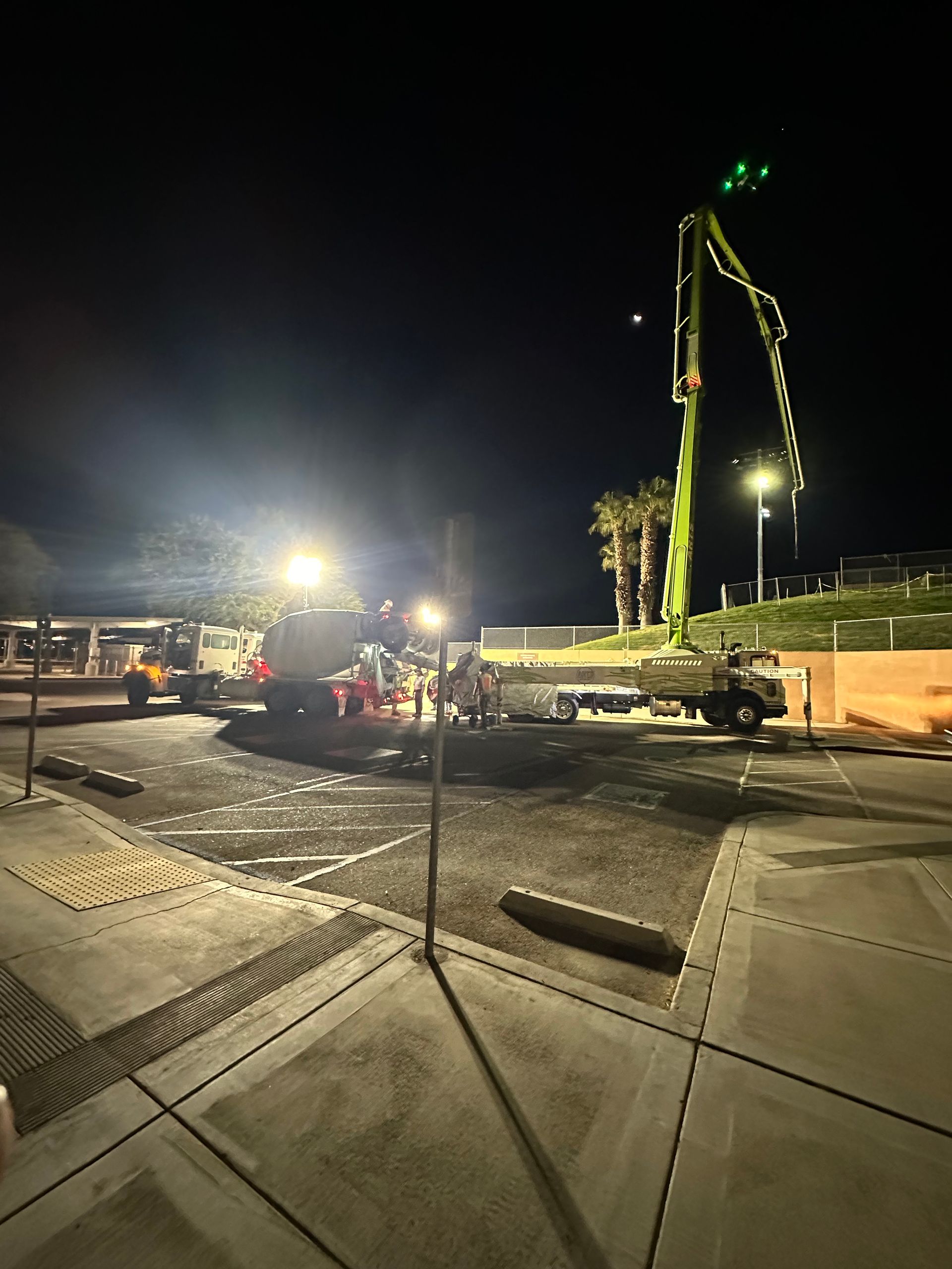 At night, a concrete pump truck and mixing truck work in a parking lot, extending their boom arm over a construction site.