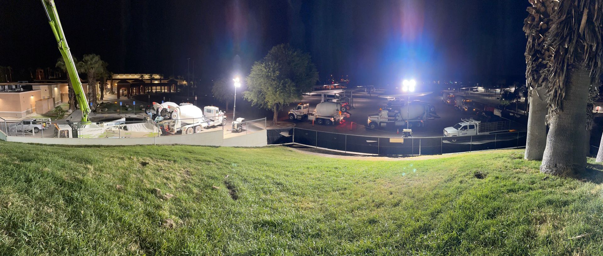 A nighttime construction site with parked trucks and heavy machinery illuminated by bright lights above a grassy hill.