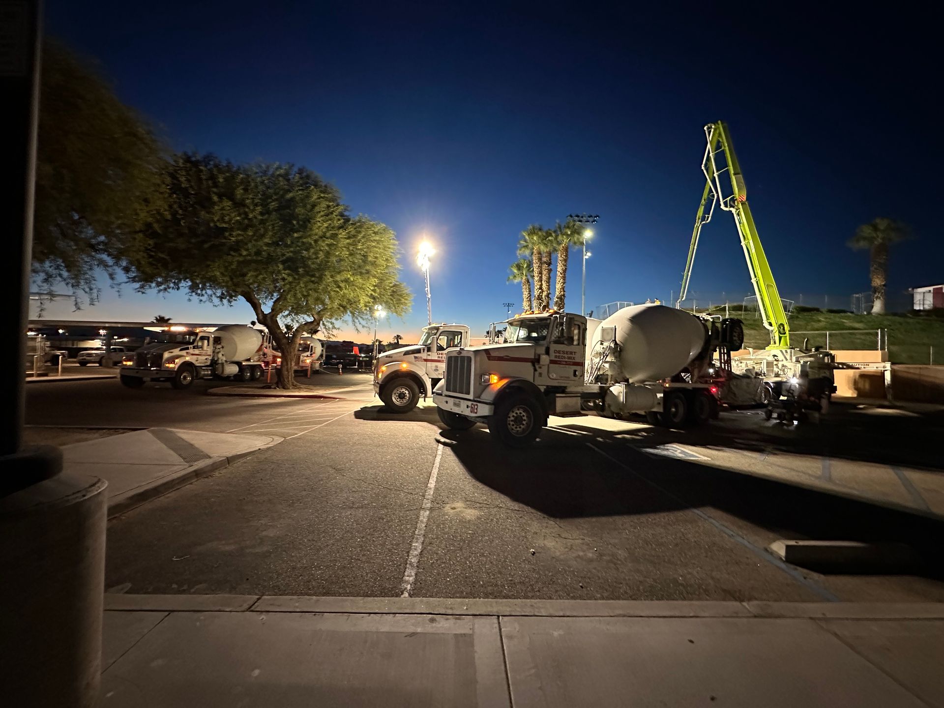 Two white concrete mixer trucks parked in a lot at dusk, with one extended mechanical boom reaching into the night sky.