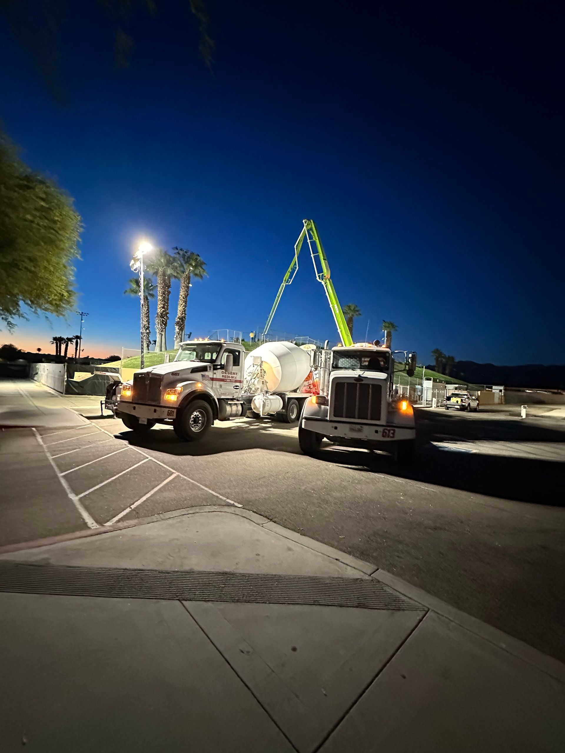 Two industrial trucks at night, one with a green concrete boom pump, parked in a paved lot under an illuminated light pole.