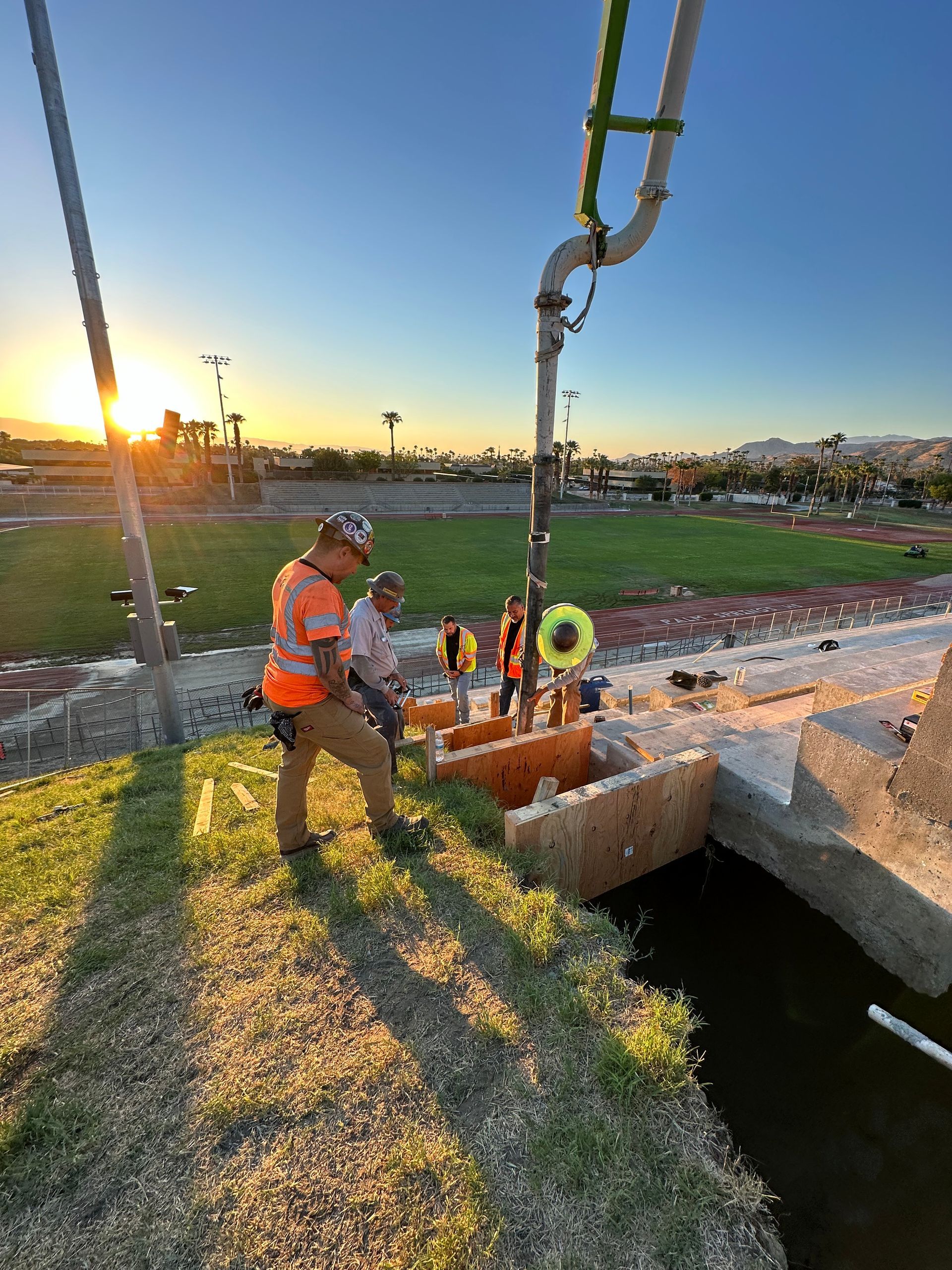 Construction crew pours concrete into wooden forms at a stadium track during sunrise.