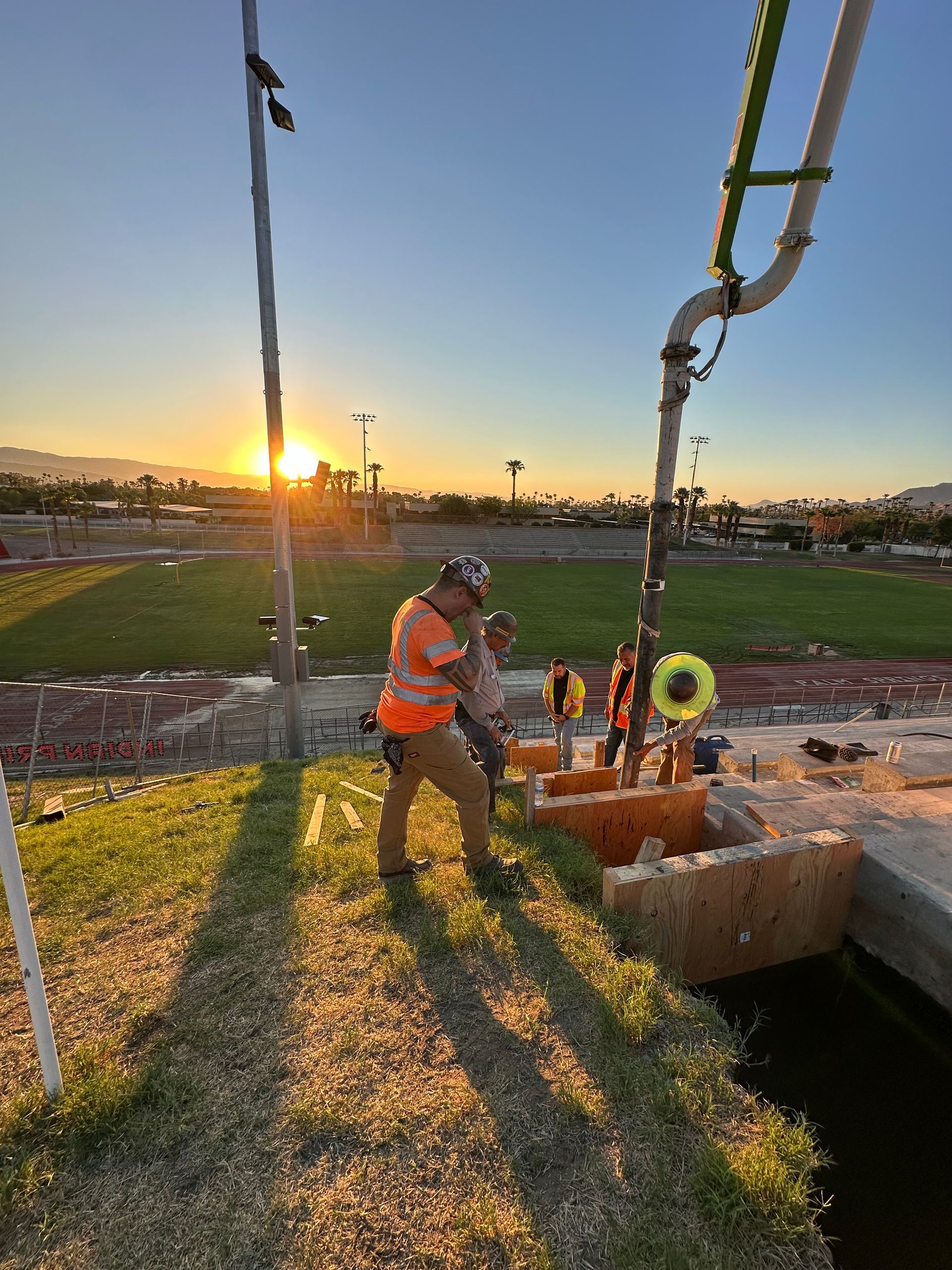 Construction crew pours concrete from a pump truck near a football field at sunrise.