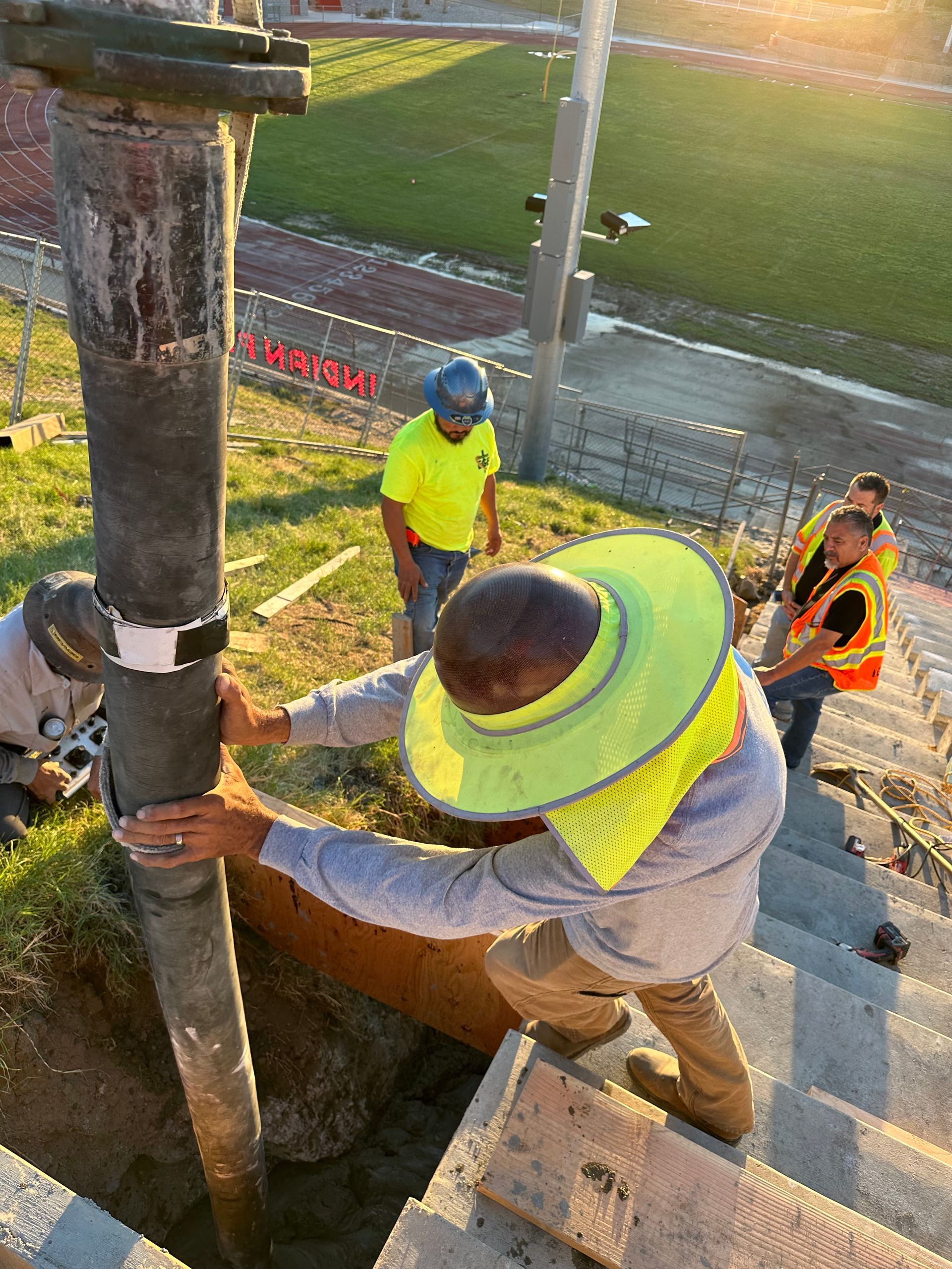 Construction workers, wearing high-visibility gear, operate a concrete pump hose at an outdoor stadium construction site.