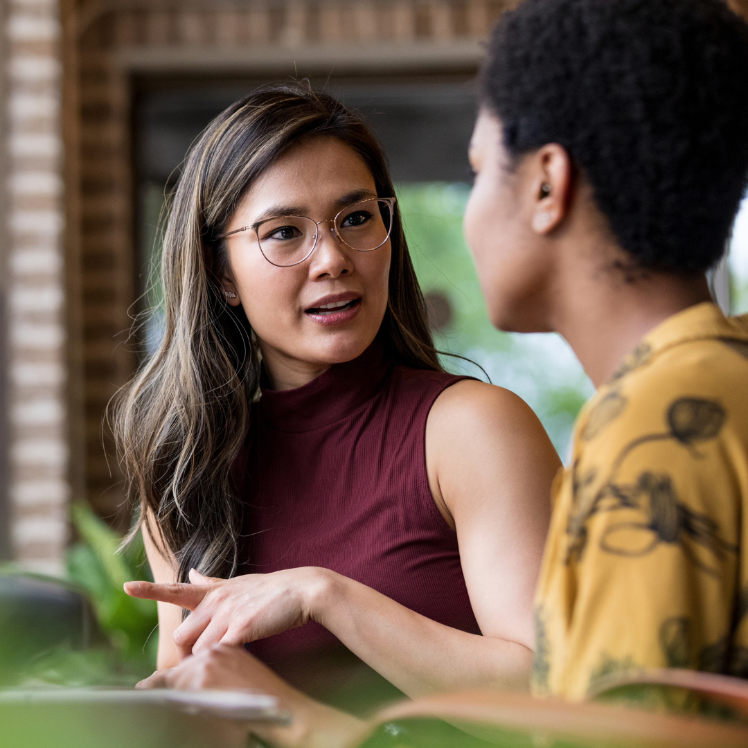 Two people having a discussion; one wears a maroon sleeveless top and glasses, gesturing while speaking to the other.